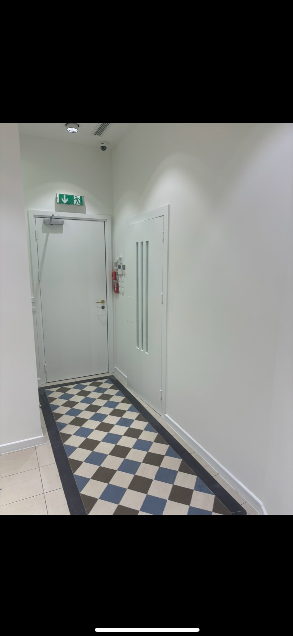Białe wnętrze korytarza z wzorzystą podłogą w szachownicę (brąz, błękit, biel), białe ściany i drzwi. Widoczny fragment sufitu z oświetleniem i kamerą.
