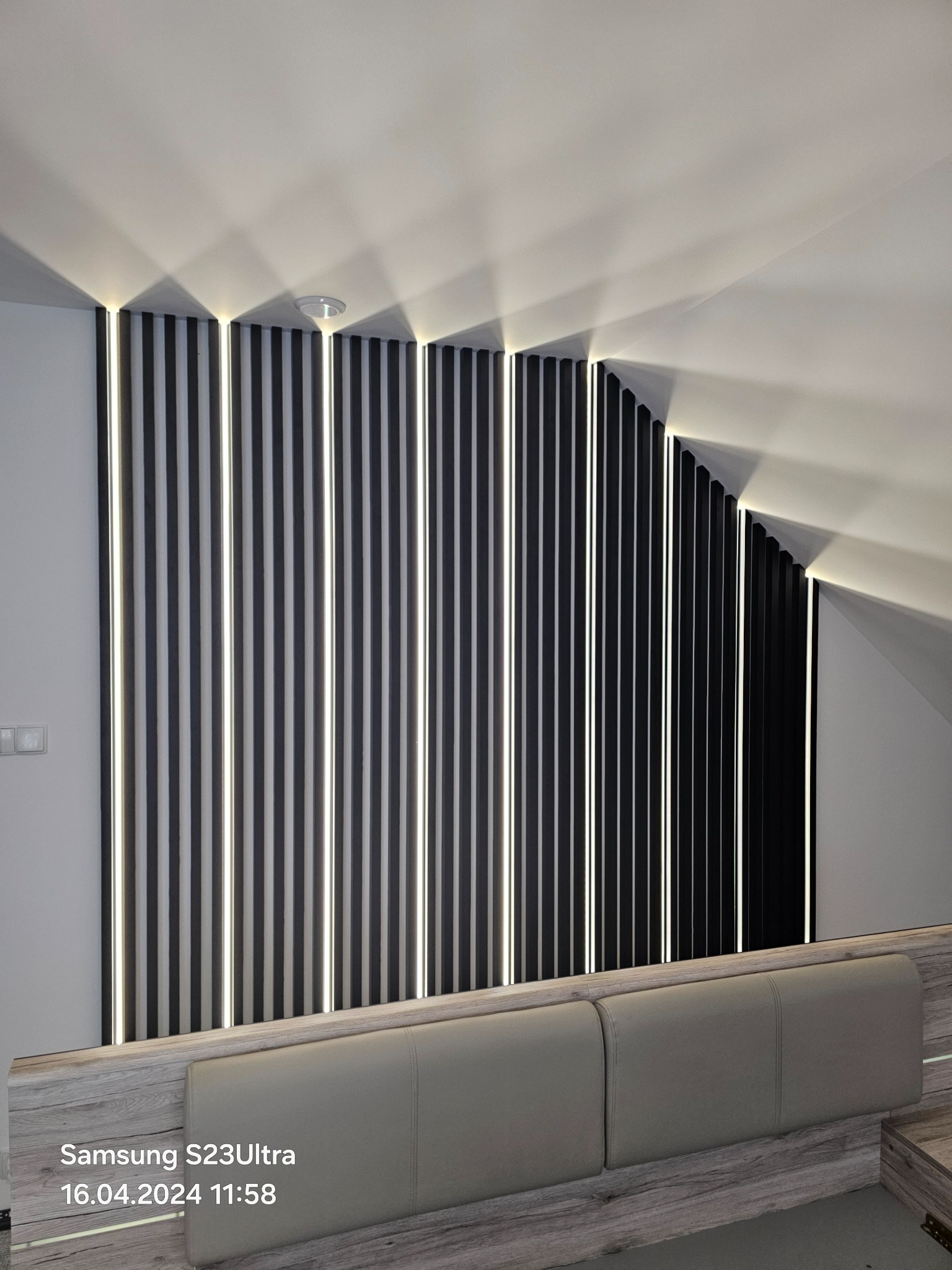 Nowoczesna ściana z czarnych paneli z wbudowanym oświetleniem LED w skosie poddasza, widoczny fragment łóżka. Minimalistyczny design wnętrza.