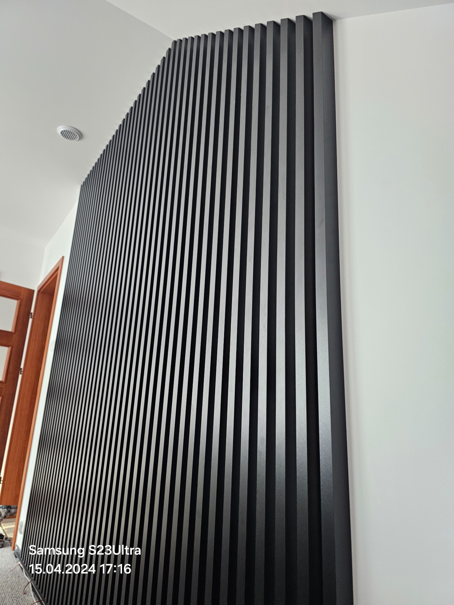 Nowoczesna ściana z czarnych, pionowych paneli dekoracyjnych, tworzących rytmiczny wzór. Minimalistyczny design, białe ściany w tle, widoczny fragment drzwi.