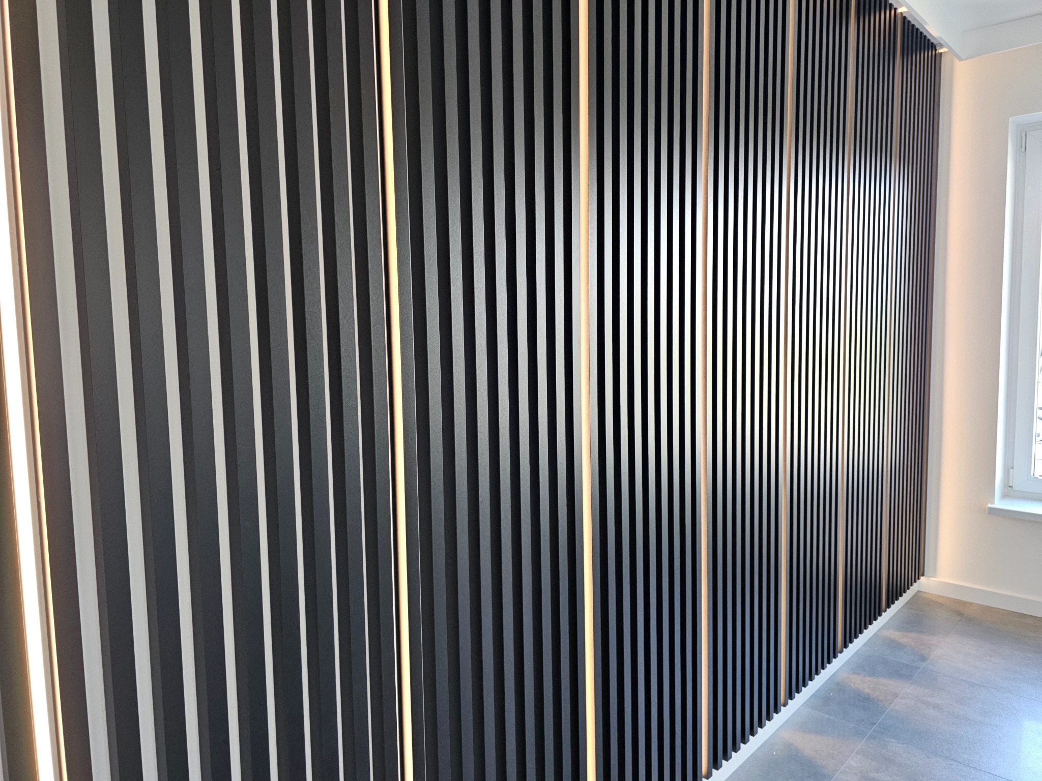 Nowoczesna ściana z pionowych, czarnych lameli z subtelnym podświetleniem LED, tworząca dekoracyjny element wnętrza. Widoczny fragment podłogi i okno.