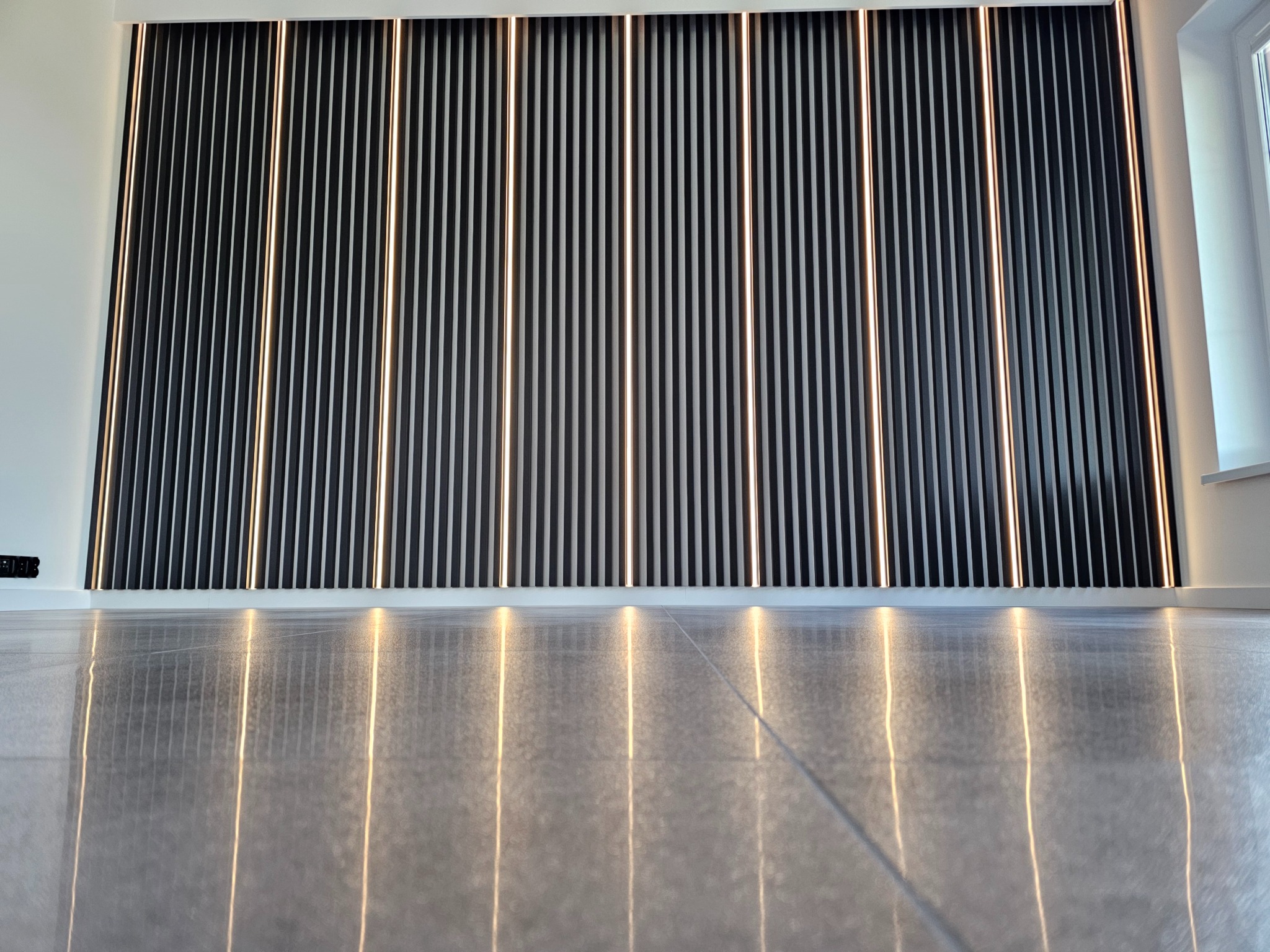 Nowoczesna ściana z czarnych lameli z podświetleniem LED, odbijająca się w polerowanej podłodze. Minimalistyczny design wnętrza z oknem po prawej stronie.