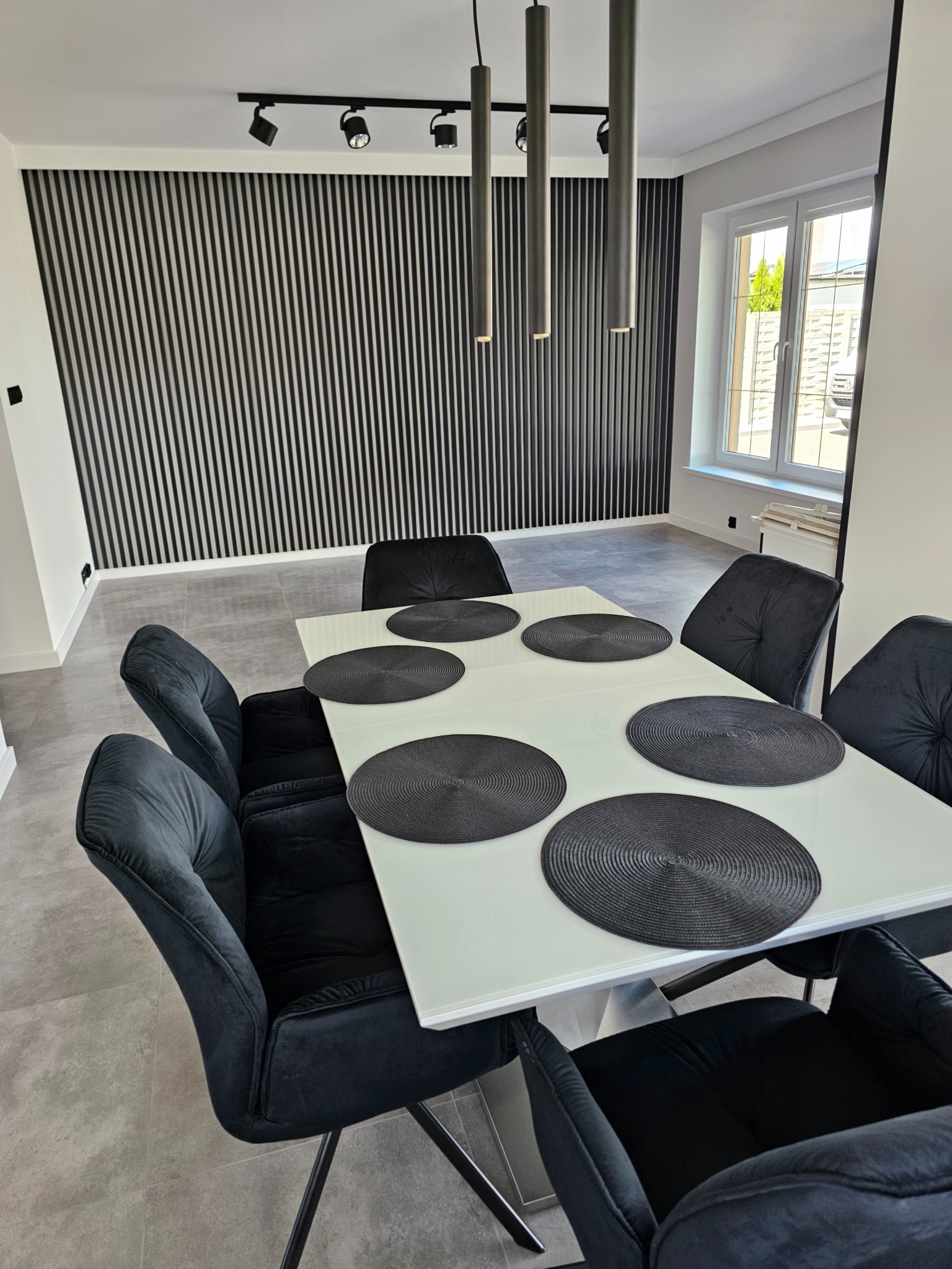 Nowoczesna jadalnia: biały stół z czarnymi podkładkami, welurowe krzesła, ściana z pionowych paneli, oświetlenie punktowe i okno z widokiem na zieleń.