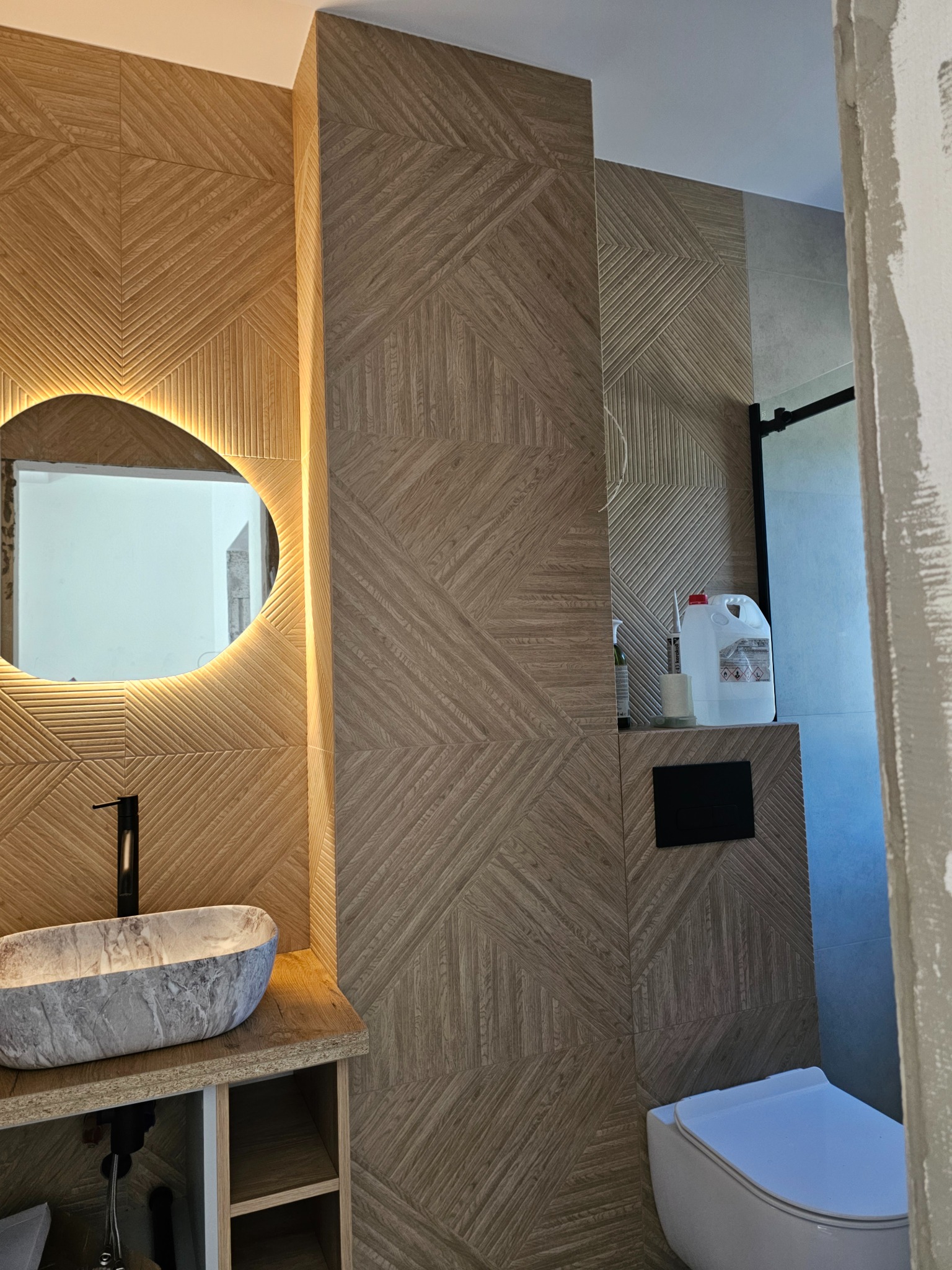 Wykończona łazienka z drewnopodobnymi płytkami ułożonymi w geometryczny wzór. Okrągłe lustro z podświetleniem LED nad umywalką z kamienia.