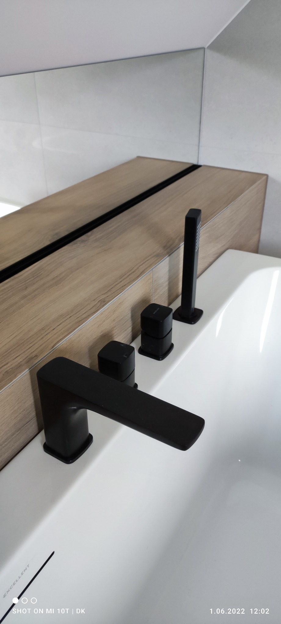 Nowoczesna armatura łazienkowa: czarna bateria umywalkowa z czterema elementami na białej umywalce, drewnopodobna szafka z lustrem w tle, minimalistyczny design.