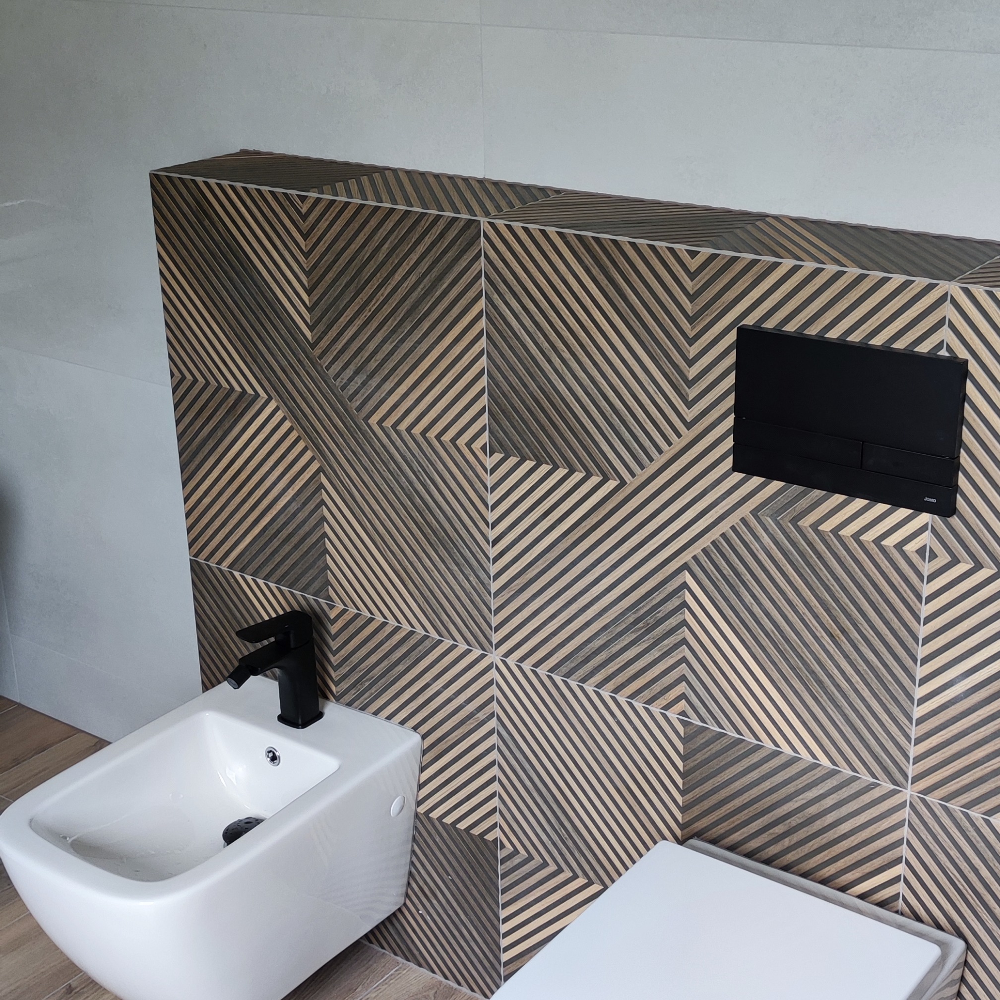 Nowoczesna łazienka z bidetem i toaletą. Dekoracyjna ściana z płytek w geometryczny wzór. Czarna armatura i przycisk spłukiwania. Jasne płytki na podłodze i ścianach.