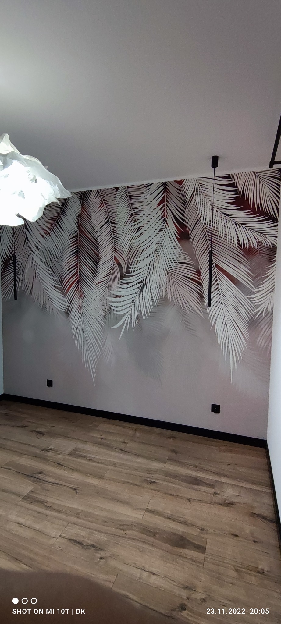 Pokój po wykończeniu: ściana z motywem liści palmowych, drewniana podłoga, nowoczesne oświetlenie sufitowe. Widok na fragment wnętrza po dekoracji.