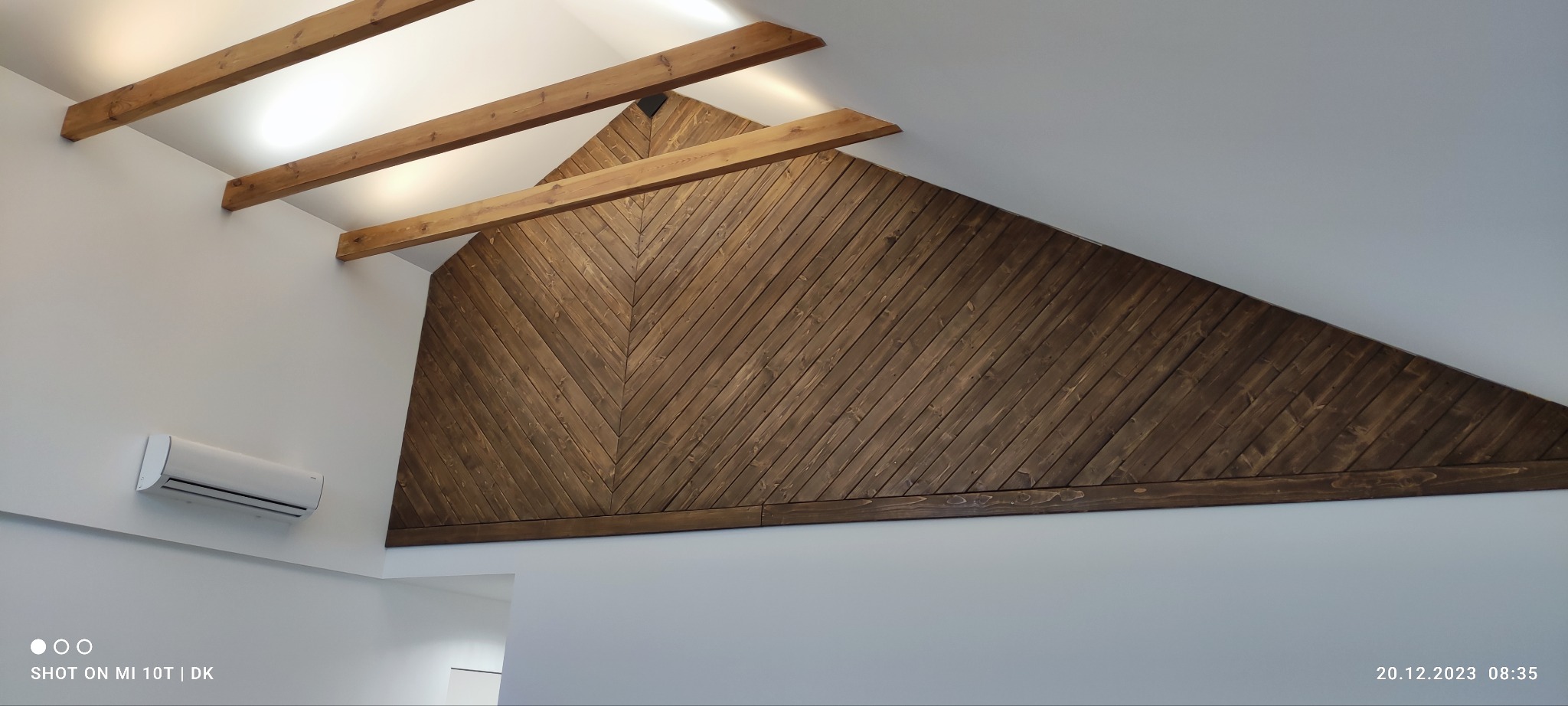 Drewniana ściana w kształcie trójkąta z desek ułożonych pod kątem, widoczne belki stropowe i klimatyzator na białej ścianie. Nowoczesne wykończenie wnętrza.