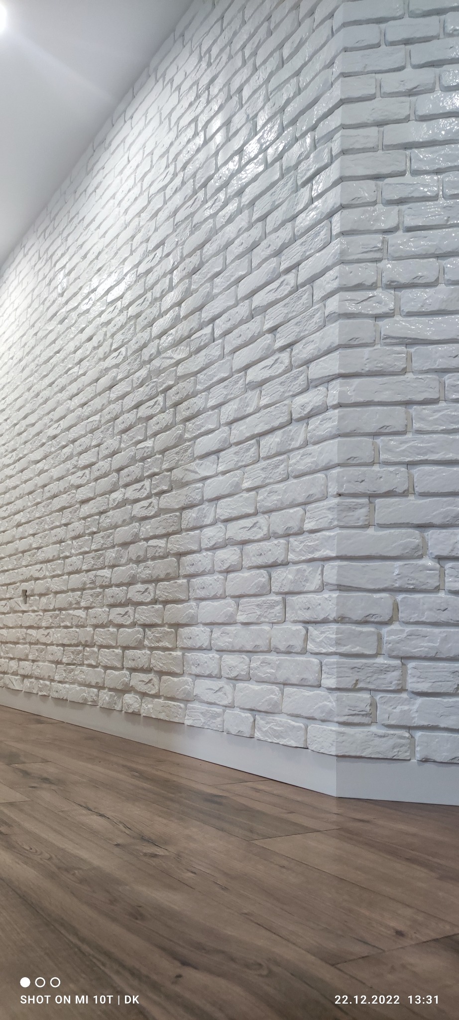 Biała ściana z cegły dekoracyjnej, widok z dołu, z delikatnym połyskiem, brązowa podłoga, listwa przypodłogowa. Nowoczesne wykończenie wnętrza.