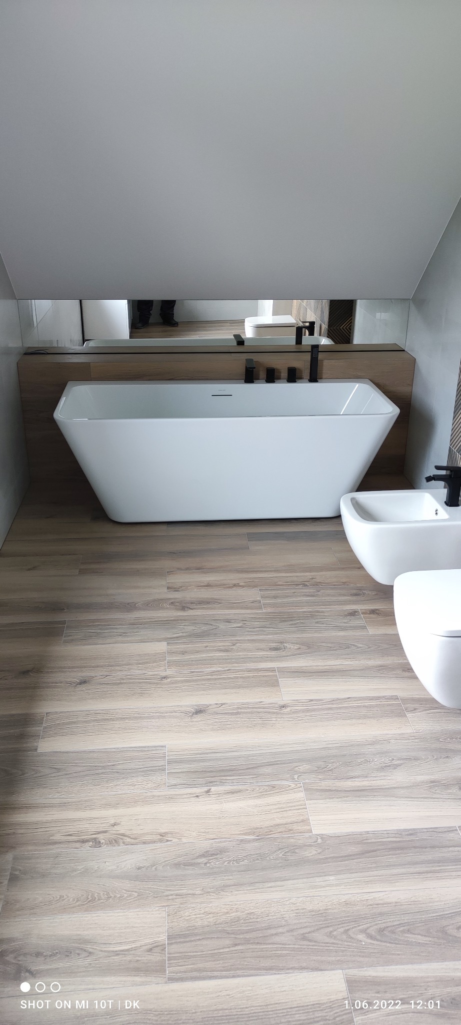 Nowoczesna łazienka na poddaszu z wolnostojącą wanną, bidetem i toaletą. Podłoga wyłożona płytkami imitującymi drewno. Minimalistyczna armatura w kolorze czarnym.