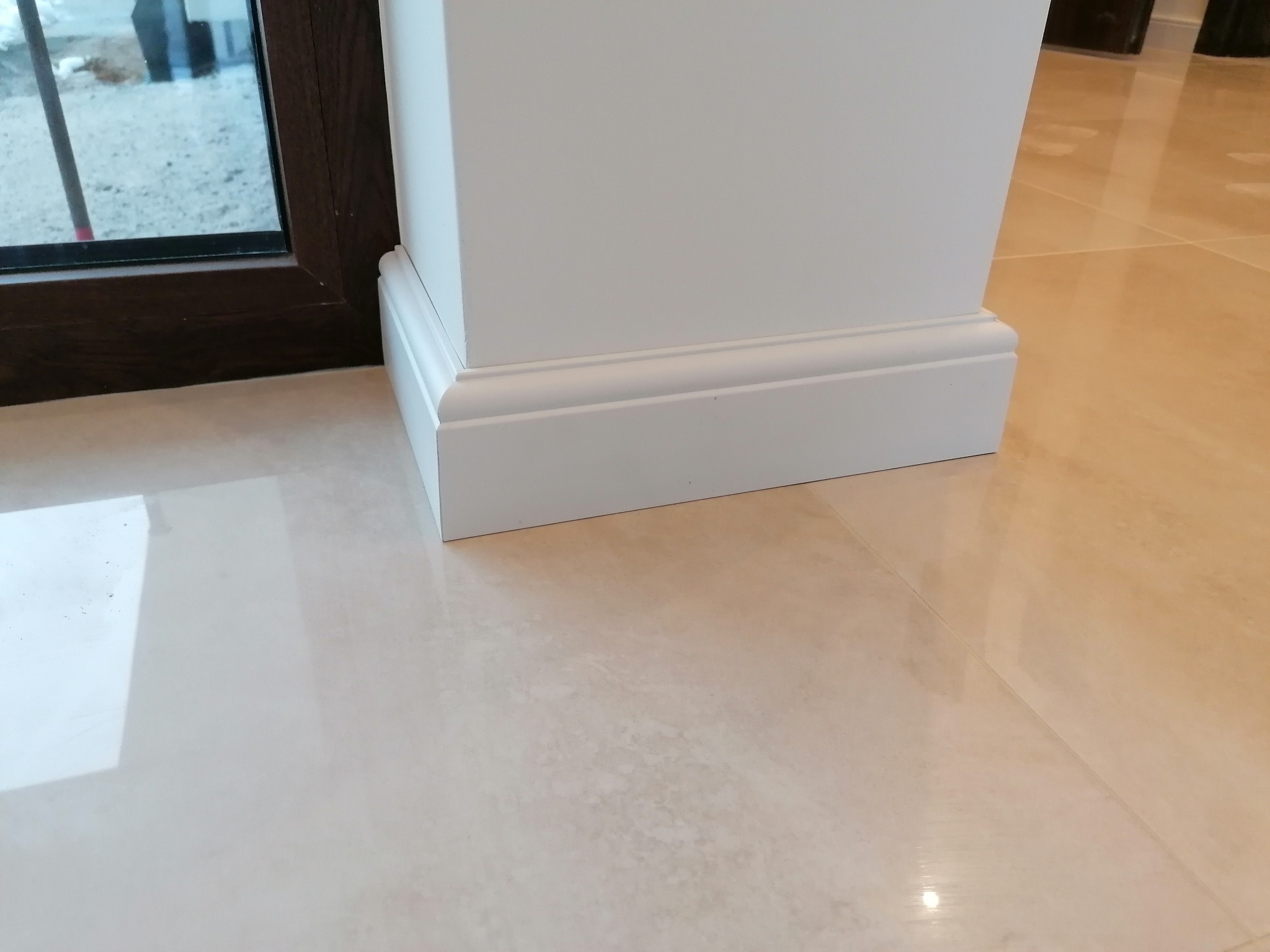 Biała listwa przypodłogowa przy ścianie i brązowej ramie okiennej na błyszczącej, beżowej podłodze z płytek. Detal wykończenia wnętrza.