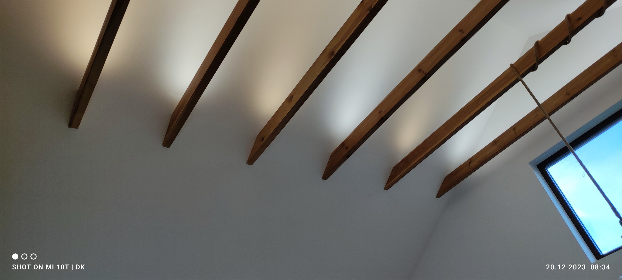 Biały sufit z drewnianymi belkami stropowymi, widok z dołu pod kątem. W tle okno dachowe z widocznym niebem. Belki w regularnych odstępach.