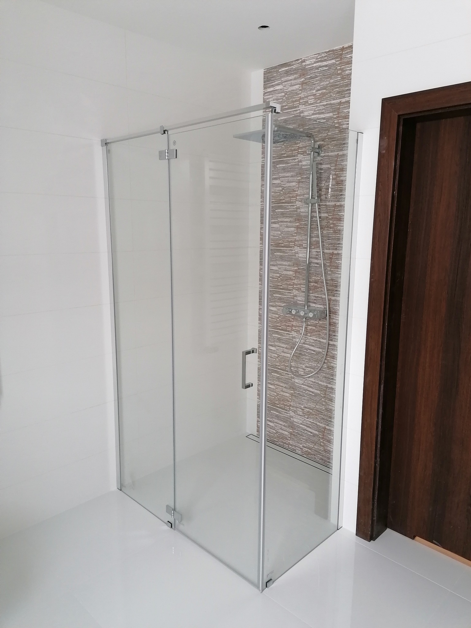 Nowoczesna kabina prysznicowa z chromowanymi okuciami i deszczownicą, na tle białych płytek i dekoracyjnej ściany z mozaiki. Ciemne, drewniane drzwi w tle.