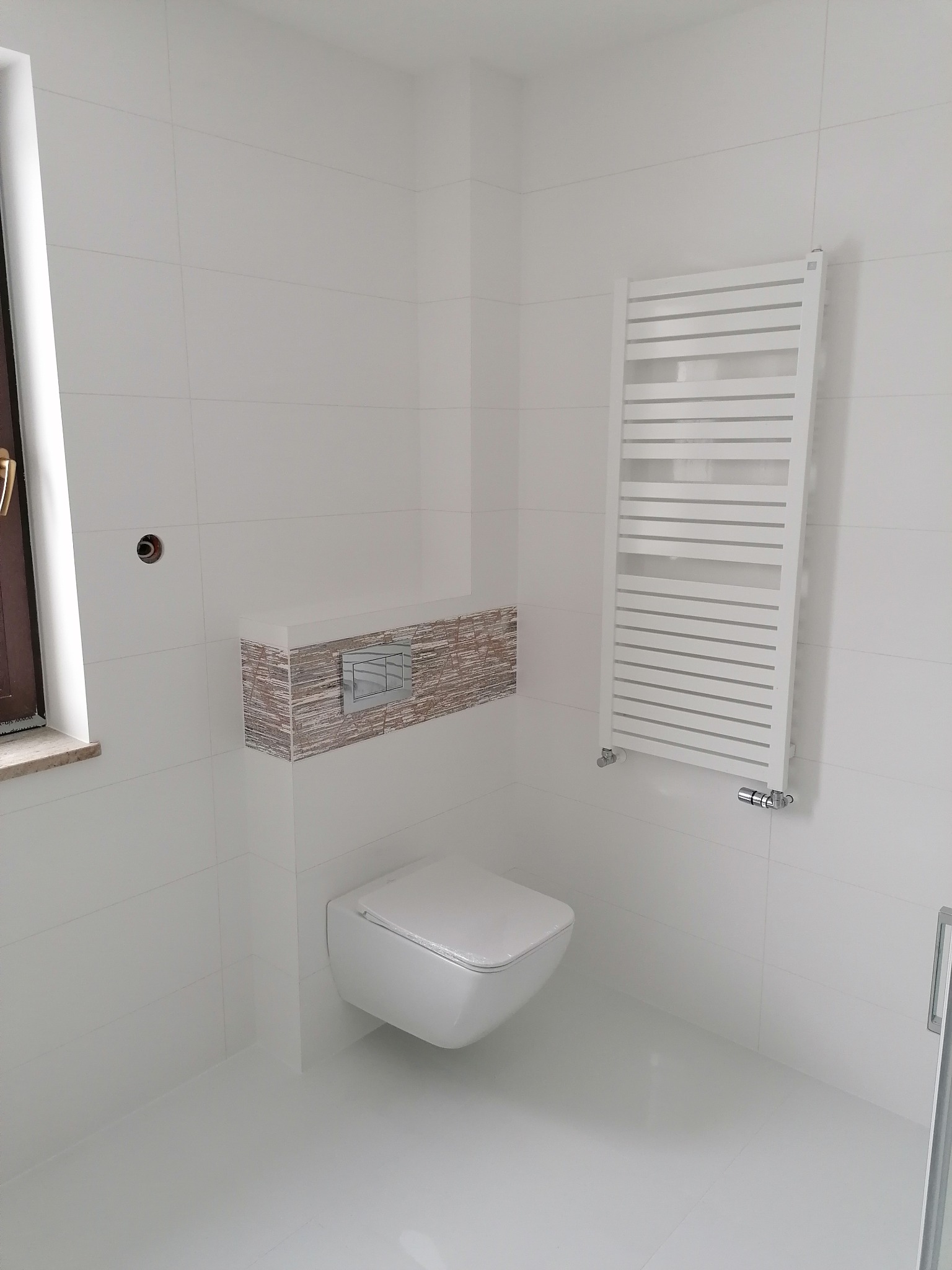 Nowoczesna, minimalistyczna łazienka z podwieszaną toaletą, grzejnikiem drabinkowym i białymi płytkami. Uwagę zwraca obudowa stelaża WC z dekoracyjną okładziną.