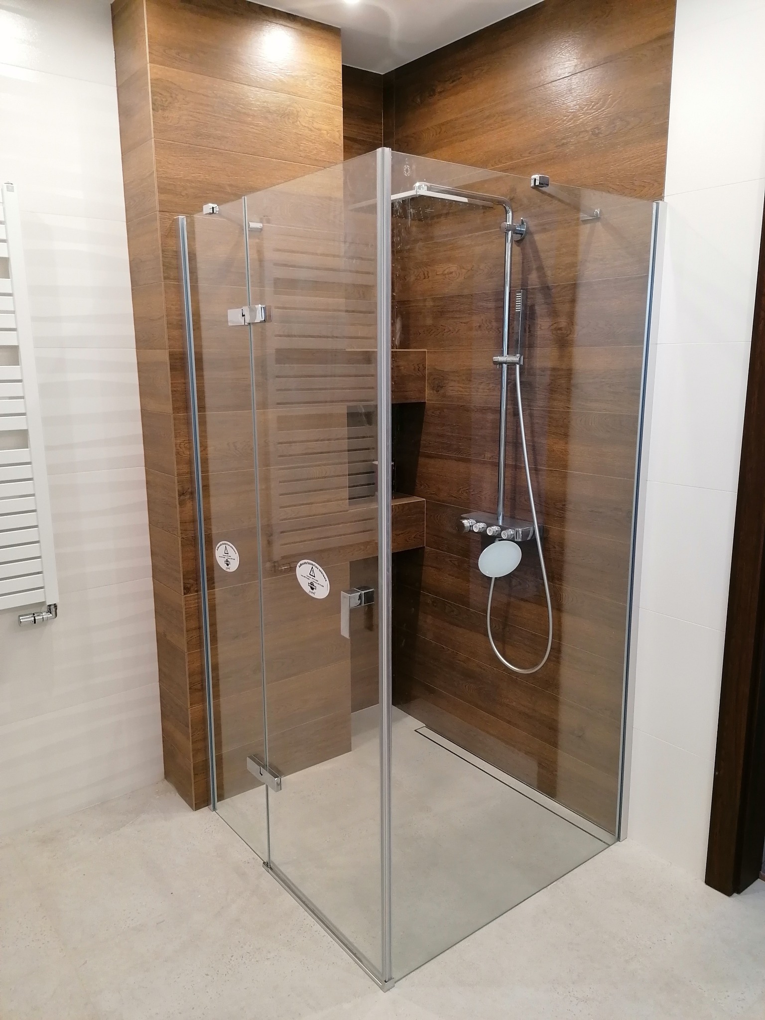 Nowoczesna kabina prysznicowa z drewnopodobnymi płytkami na ścianach i szklanymi drzwiami, chromowana armatura, jasna podłoga. Minimalistyczny design łazienki.