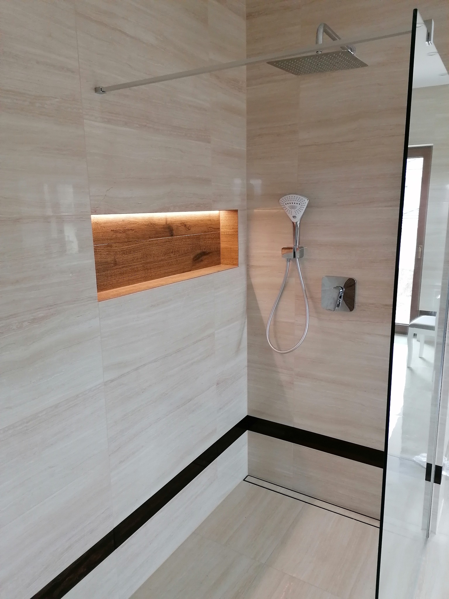 Nowoczesna kabina prysznicowa z beżowymi płytkami imitującymi marmur, drewnianą wnęką z oświetleniem i czarnym akcentem w postaci odpływu liniowego.