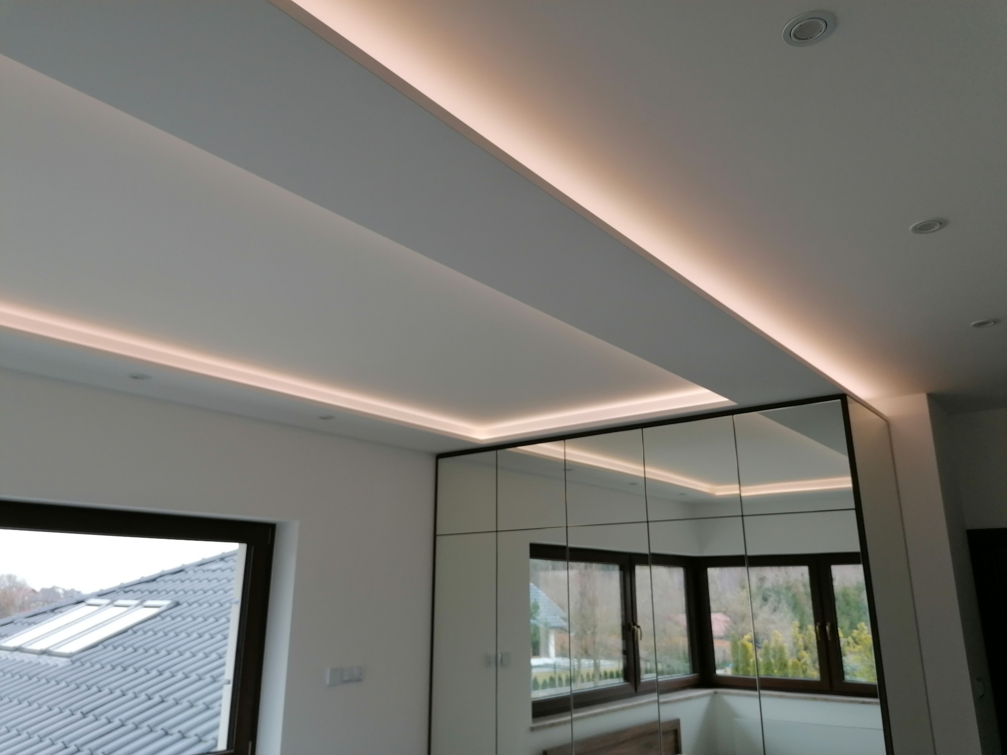 Białe wnętrze z podwieszanym sufitem i oświetleniem LED. Lustrzana szafa w zabudowie odbija okno. Minimalistyczny design.