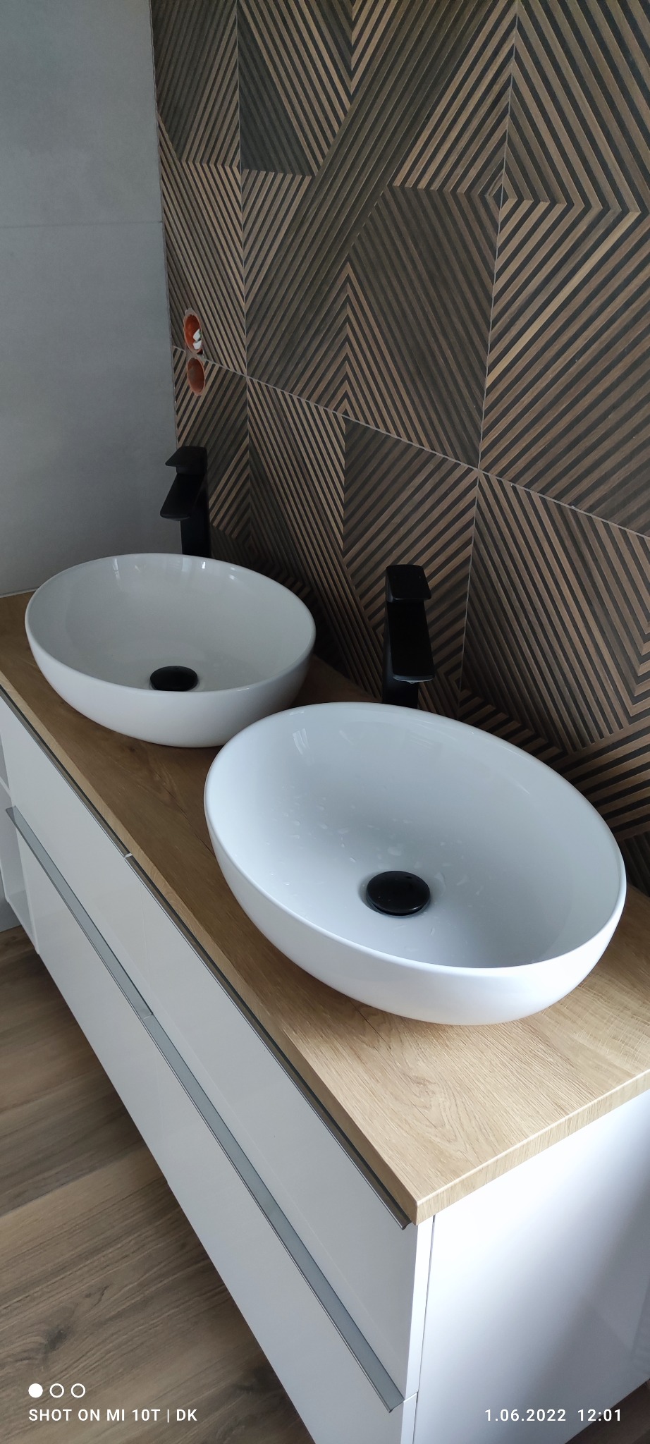 Dwie białe, owalne umywalki nablatowe z czarnymi bateriami na drewnianym blacie. W tle dekoracyjna ściana z geometrycznym wzorem. Nowoczesna aranżacja łazienki.