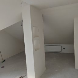 entreprise - Wnętrze poddasza po szpachlowaniu i malowaniu na biało. Widoczne wnęki w ścianie, grzejnik i fragmenty podłogi. Minimalistyczny design, surowe wykończenie.