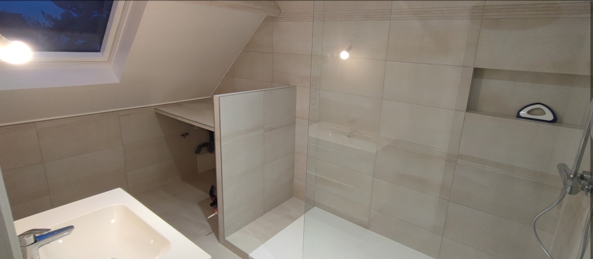 Jasna łazienka na poddaszu z beżowymi płytkami, kabiną prysznicową ze szklaną ścianką i umywalką. Widoczne okno dachowe i półka w ścianie.