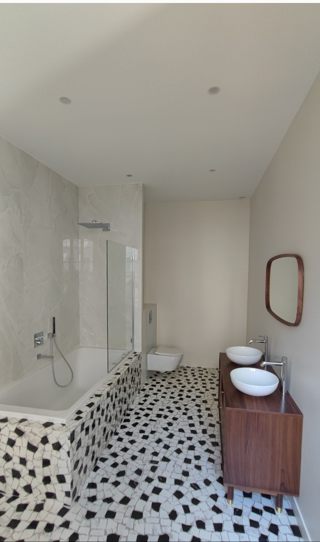 Nowoczesna łazienka z wanną obudowaną mozaiką, podwójną umywalką na drewnianej szafce i lustrem o zaokrąglonym kształcie. Białe ściany i mozaikowa podłoga.