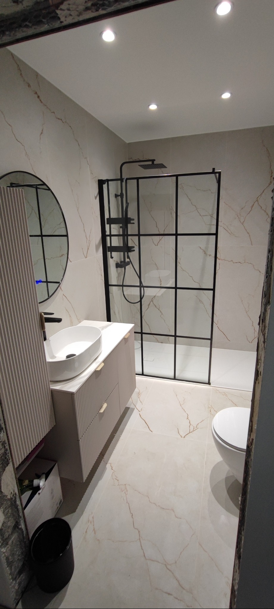 Wykończona łazienka z kabiną typu walk-in, umywalką nablatową i okrągłym lustrem. Płytki imitujące marmur na ścianach i podłodze. Nowoczesny design.