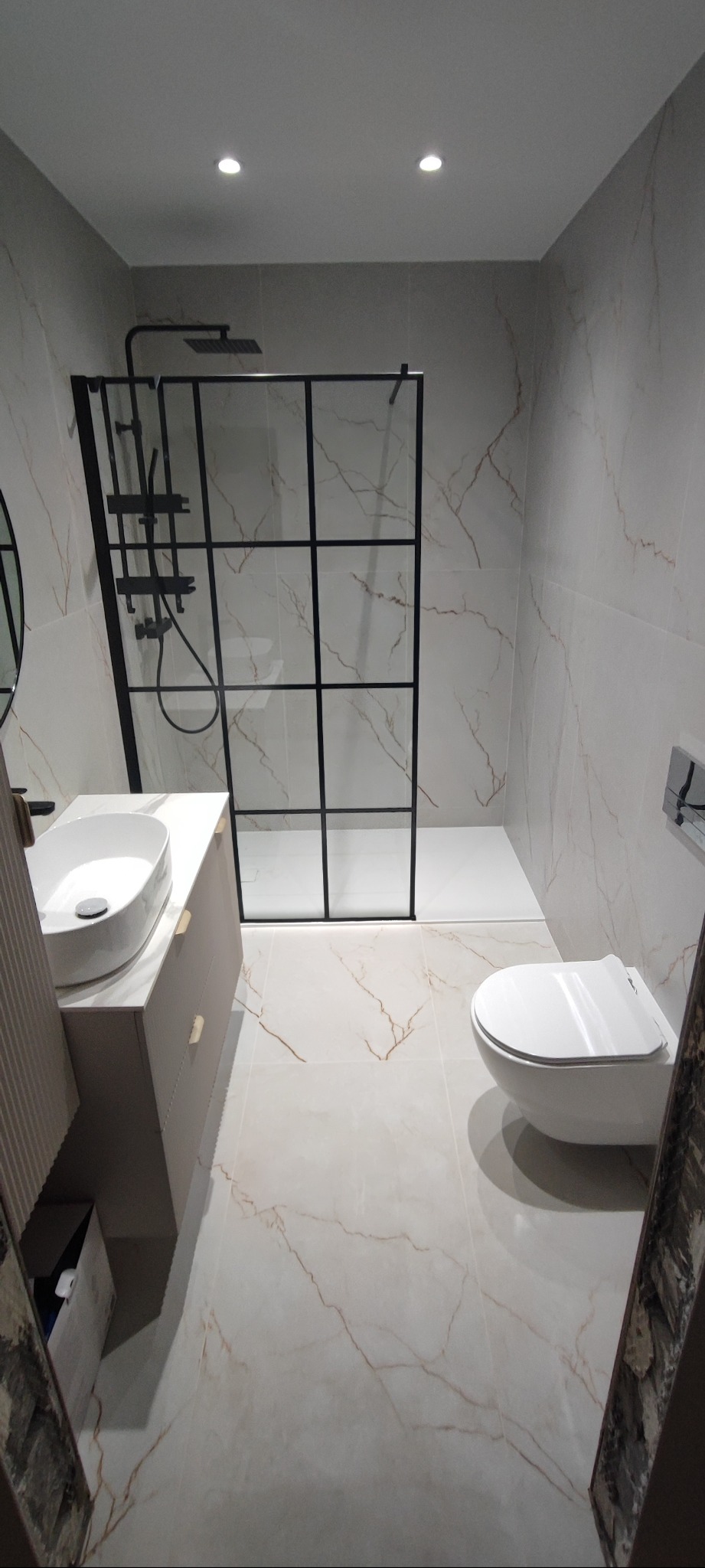 Nowoczesna łazienka z kabiną prysznicową typu walk-in z czarnymi profilami, umywalką nablatową i podwieszaną toaletą. Płytki imitujące marmur na ścianach i podłodze.