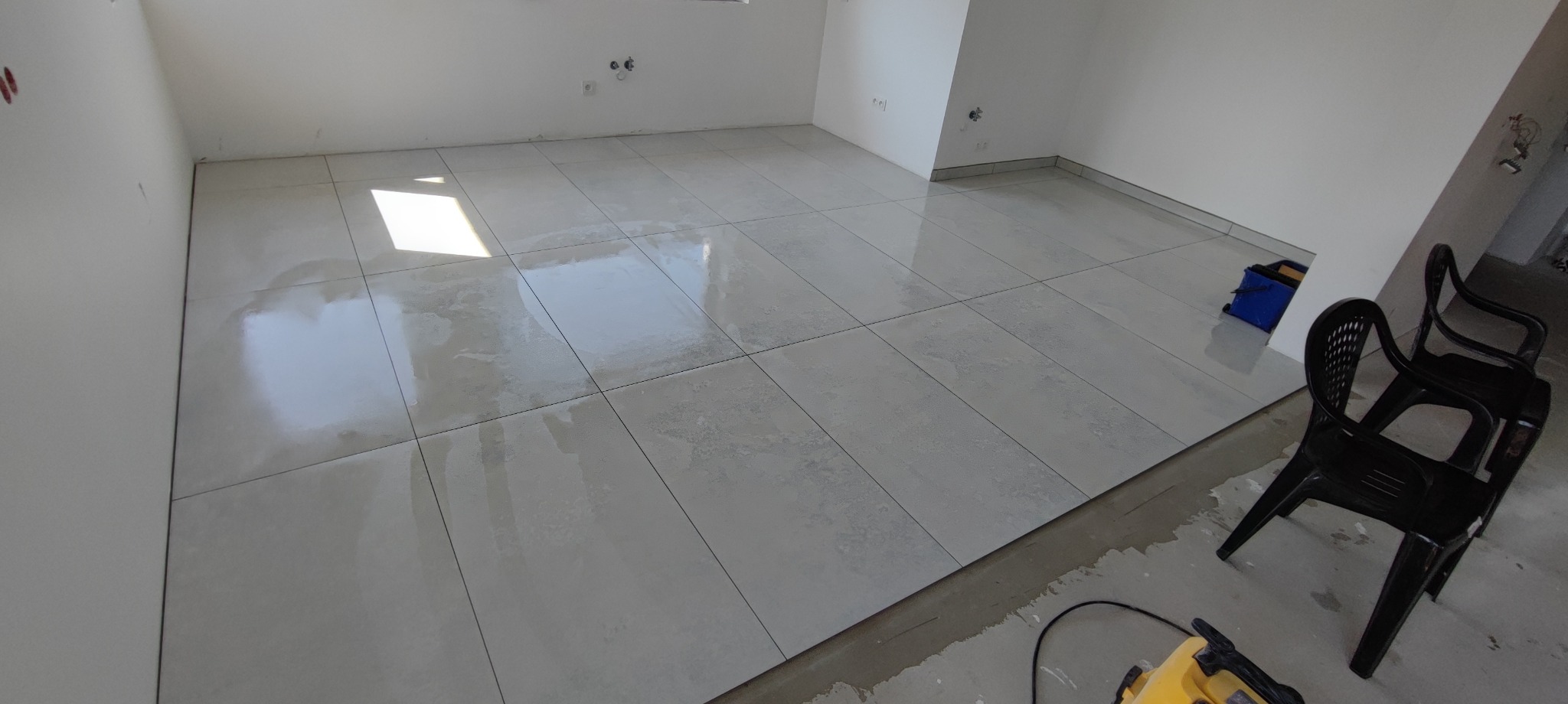 Jasne, świeżo ułożone płytki podłogowe w pustym, jasnym pomieszczeniu. Widoczne narzędzia glazurnicze i krzesła. Minimalistyczne wnętrze po remoncie.