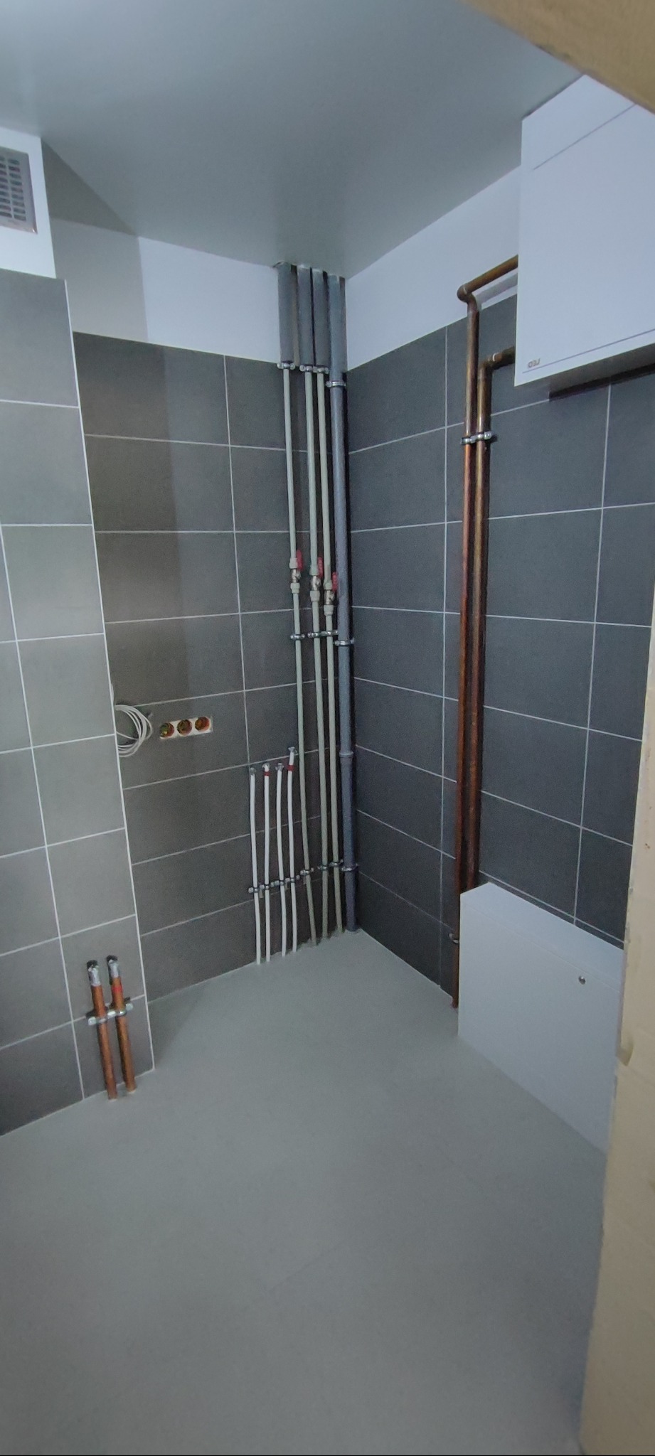 Szare płytki w łazience z widoczną instalacją wodną i grzewczą. Rury miedziane i plastikowe, szare fugi. Minimalistyczny design, świeżo po remoncie.