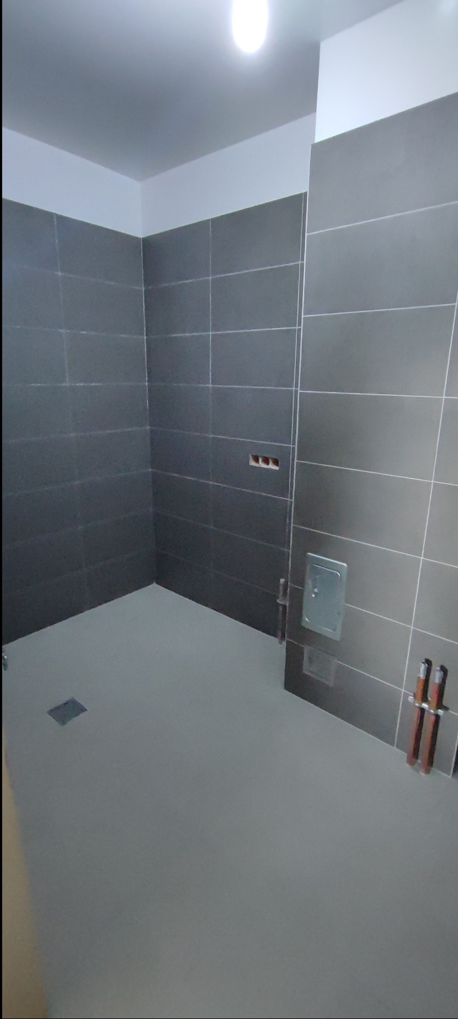 Wykończona łazienka z szarymi, kwadratowymi płytkami, widoczne przyłącza hydrauliczne i kratka ściekowa. Minimalistyczny design, surowe wykończenie.