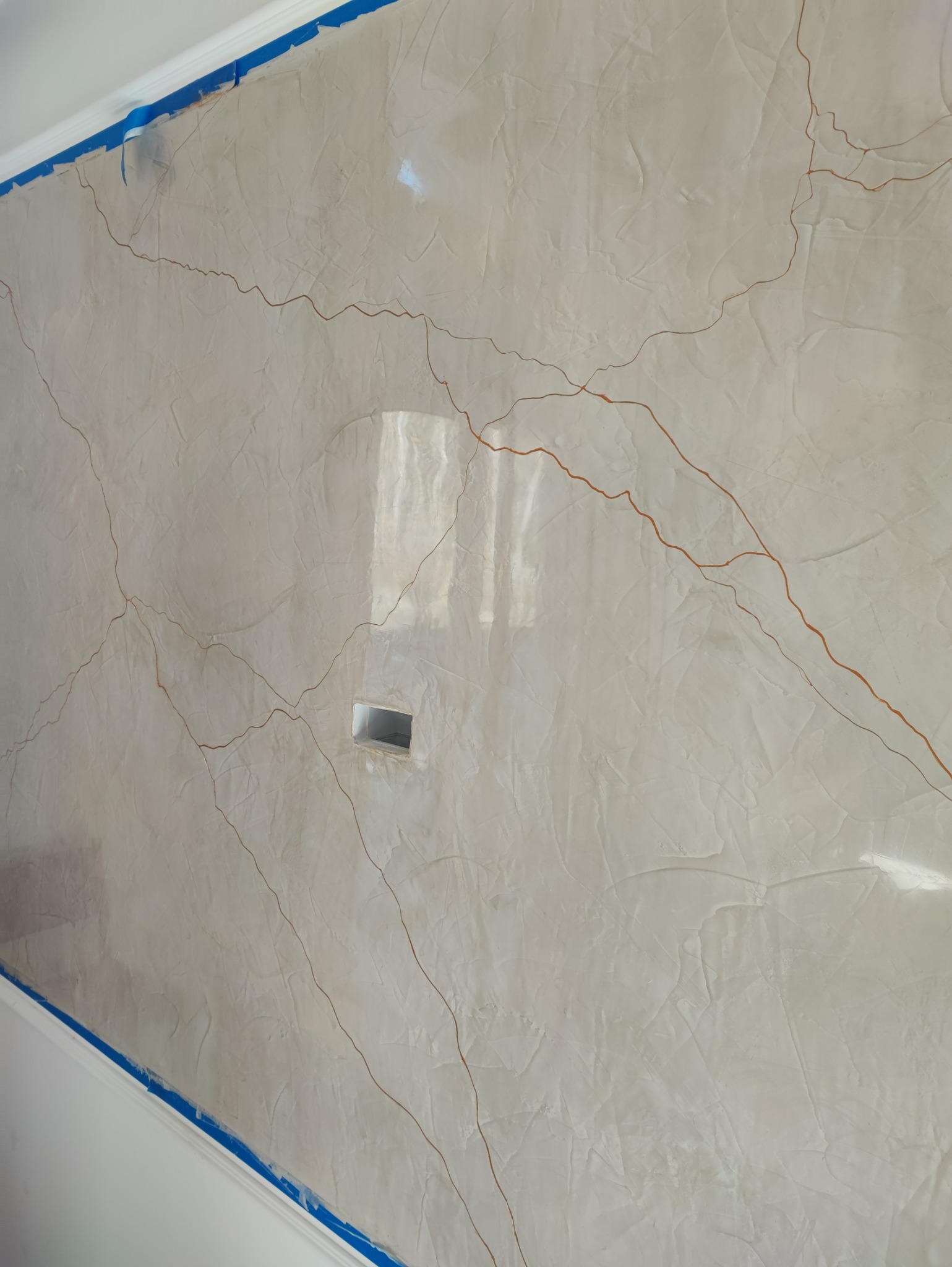 Beżowa ściana z efektem spękań, zabezpieczona niebieską taśmą malarską przy suficie i podłodze. Widoczny otwór na gniazdko. Dekoracyjne wykończenie.