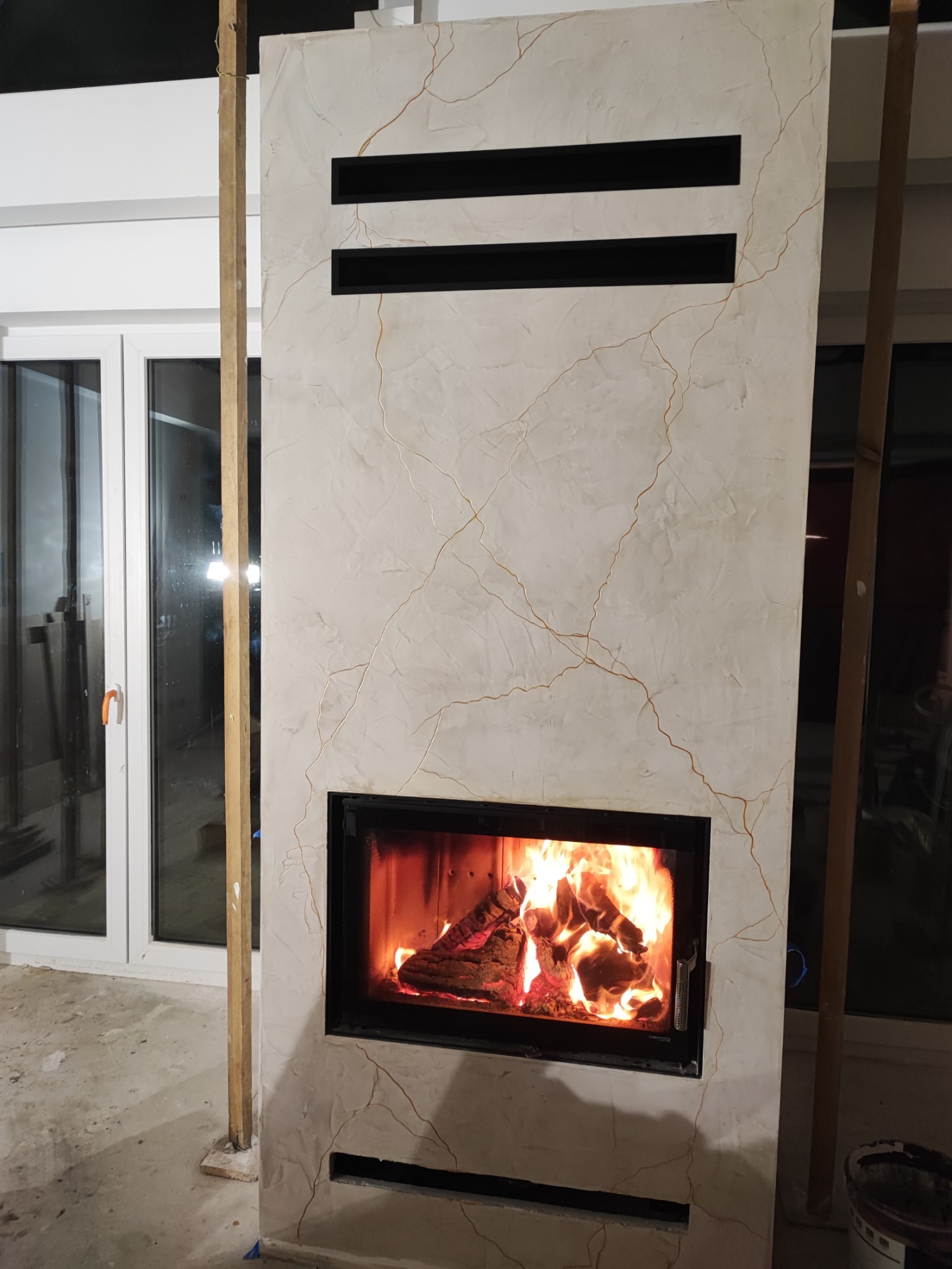 Nowoczesny kominek z marmurowym wykończeniem i paleniskiem w trakcie budowy, widoczne drewniane podpory i surowa podłoga. Detal wykończenia wnętrza.