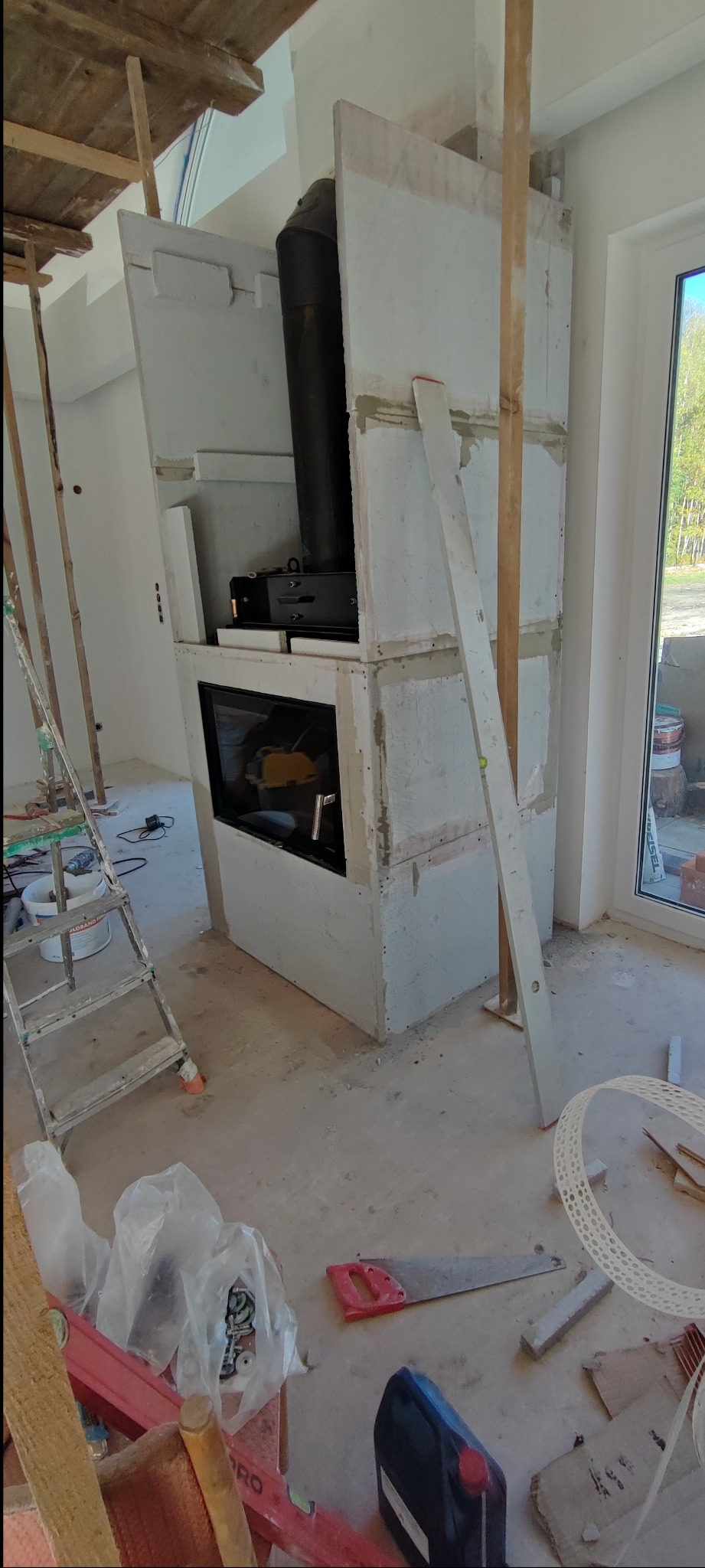 Wnętrze w trakcie budowy kominka z płyt. Widoczne rusztowania, narzędzia i materiały budowlane. Kominek w surowym stanie, z widoczną konstrukcją i wkładem kominkowym.