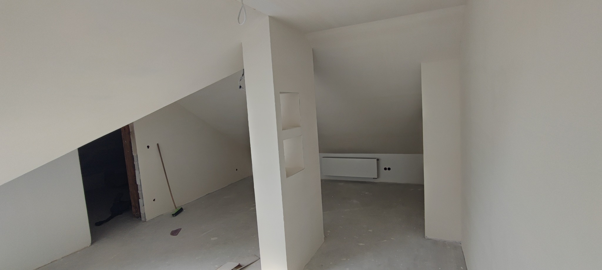 Wnętrze poddasza po szpachlowaniu i malowaniu na biało. Widoczne wnęki w ścianie, grzejnik i fragmenty podłogi. Minimalistyczny design, surowe wykończenie.
