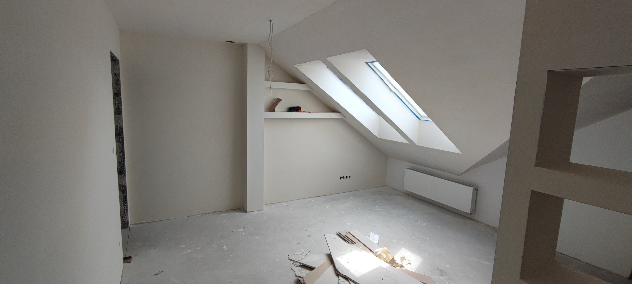 Jasne poddasze po remoncie: skośne okno dachowe, świeżo pomalowane ściany w odcieniach bieli, grzejnik, widoczne ślady po gniazdkach elektrycznych, surowe wykończenie podłogi.