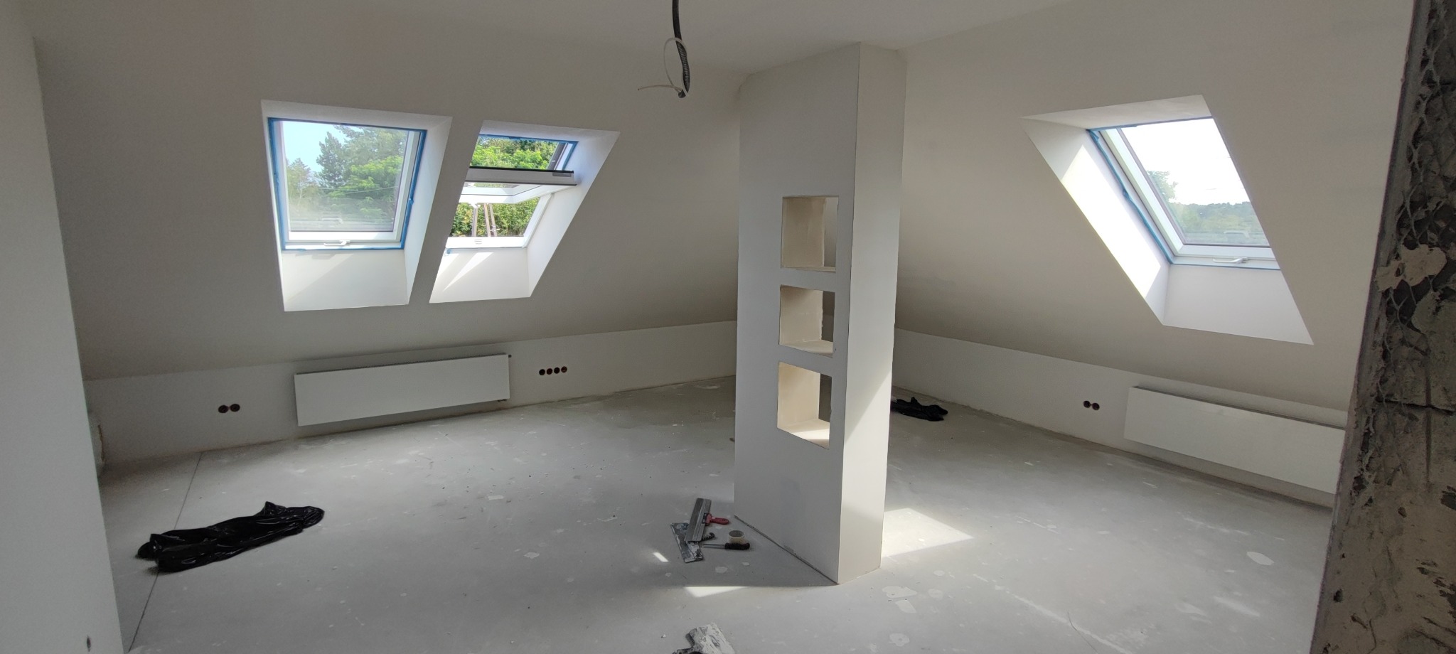 Poddasze po szpachlowaniu i malowaniu na biało, z oknami dachowymi i wnękami w ścianie. Widoczne narzędzia malarskie i folia ochronna na podłodze.