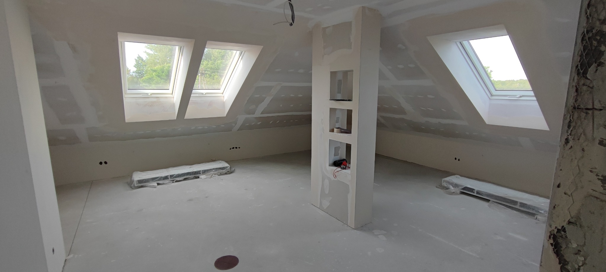Poddasze w trakcie wykończenia: ściany i skosy pokryte płytami gipsowo-kartonowymi, widoczne okna dachowe i wnęka w ścianie. Grzejniki zabezpieczone folią.