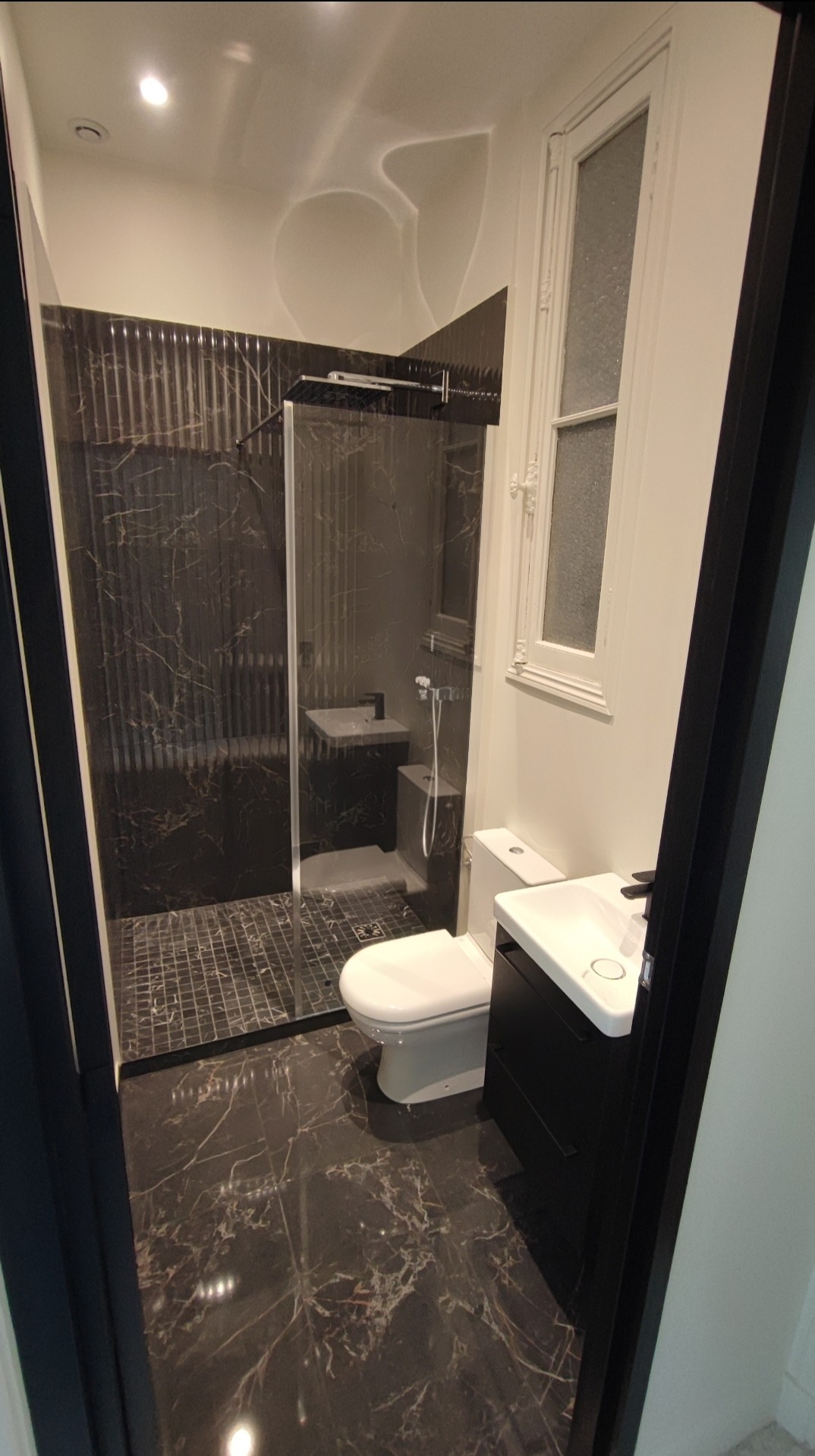 Nowoczesna łazienka z kabiną prysznicową typu walk-in, czarnymi płytkami imitującymi marmur na ścianach i podłodze, białą armaturą i oknem z ozdobną ramą.