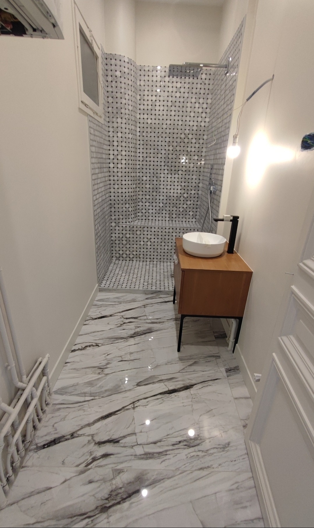 Nowoczesna łazienka z kabiną prysznicową wyłożoną mozaiką w czarno-białe kropki, marmurową podłogą i drewnianą szafką z umywalką. Minimalistyczny design.