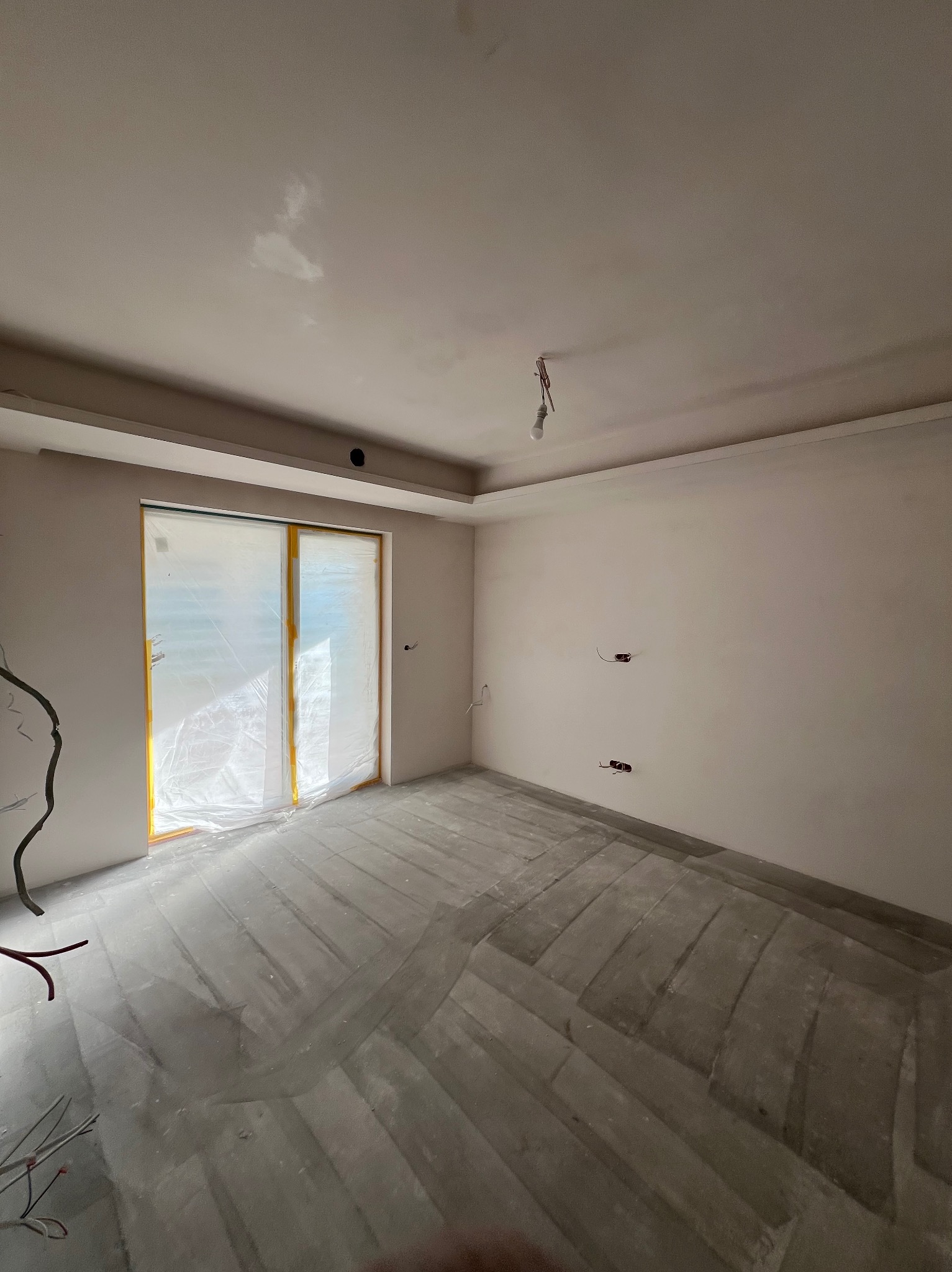 Pokój w trakcie remontu: ściany po szpachlowaniu, zabezpieczone okno folią, widoczne kable elektryczne. Podłoga przygotowana pod wylewkę.