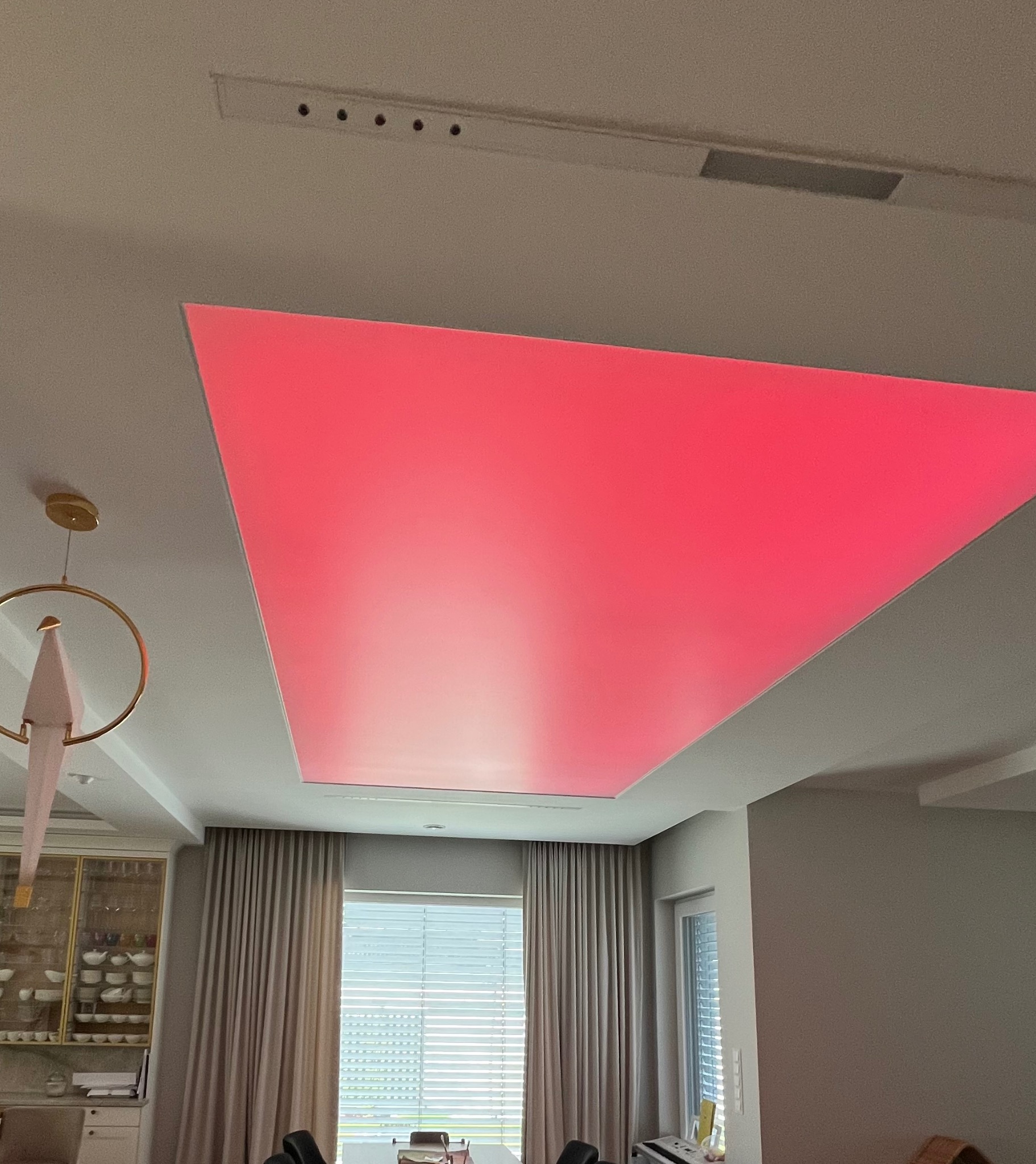 Nowoczesny sufit podwieszany z oświetleniem LED w kolorze różowym, widok z dołu, fragment żyrandola i okna z zasłonami w tle.