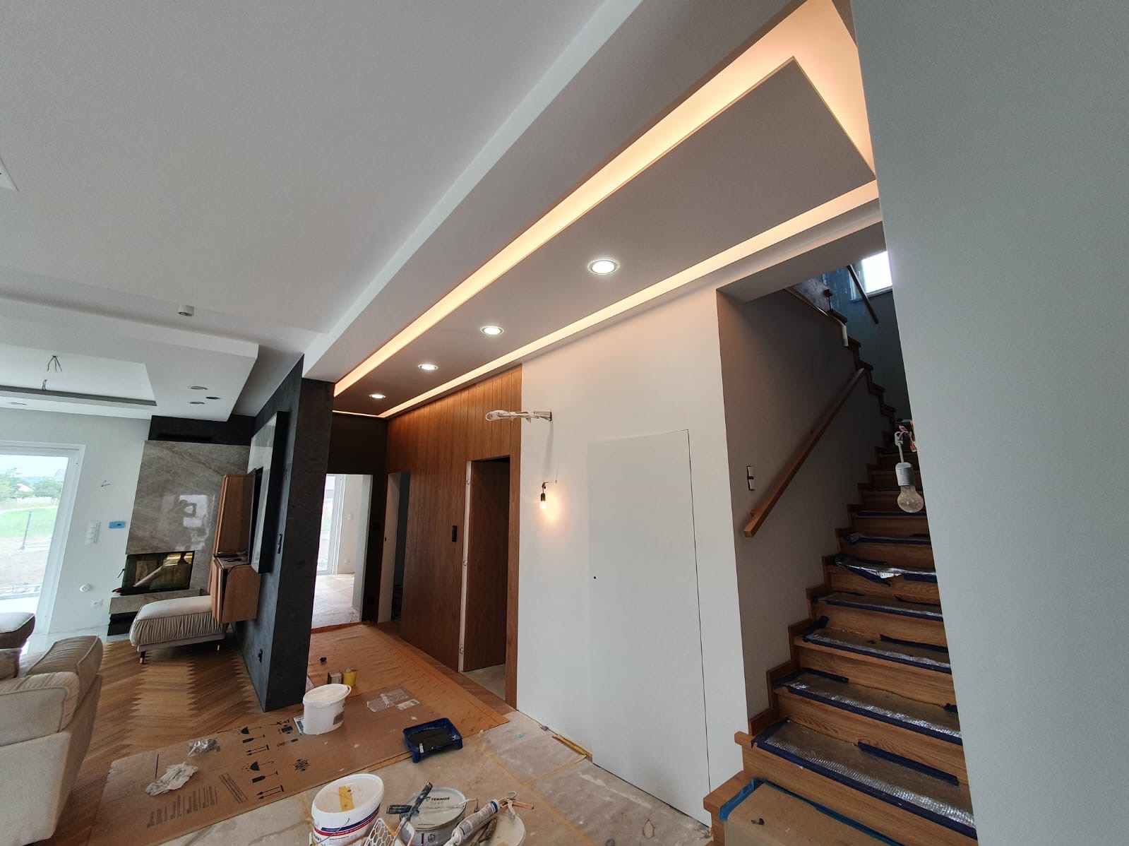 Nowoczesne wnętrze w trakcie wykańczania: białe ściany, drewniane schody zabezpieczone folią, podwieszany sufit z oświetleniem LED. Widoczne narzędzia malarskie.