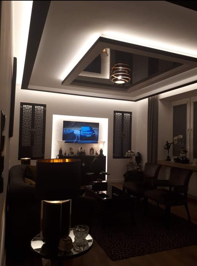 Nowoczesny salon z podwieszanym sufitem, oświetleniem LED i dekoracyjnymi panelami ściennymi. Centralnie umieszczony telewizor i designerska lampa wisząca.