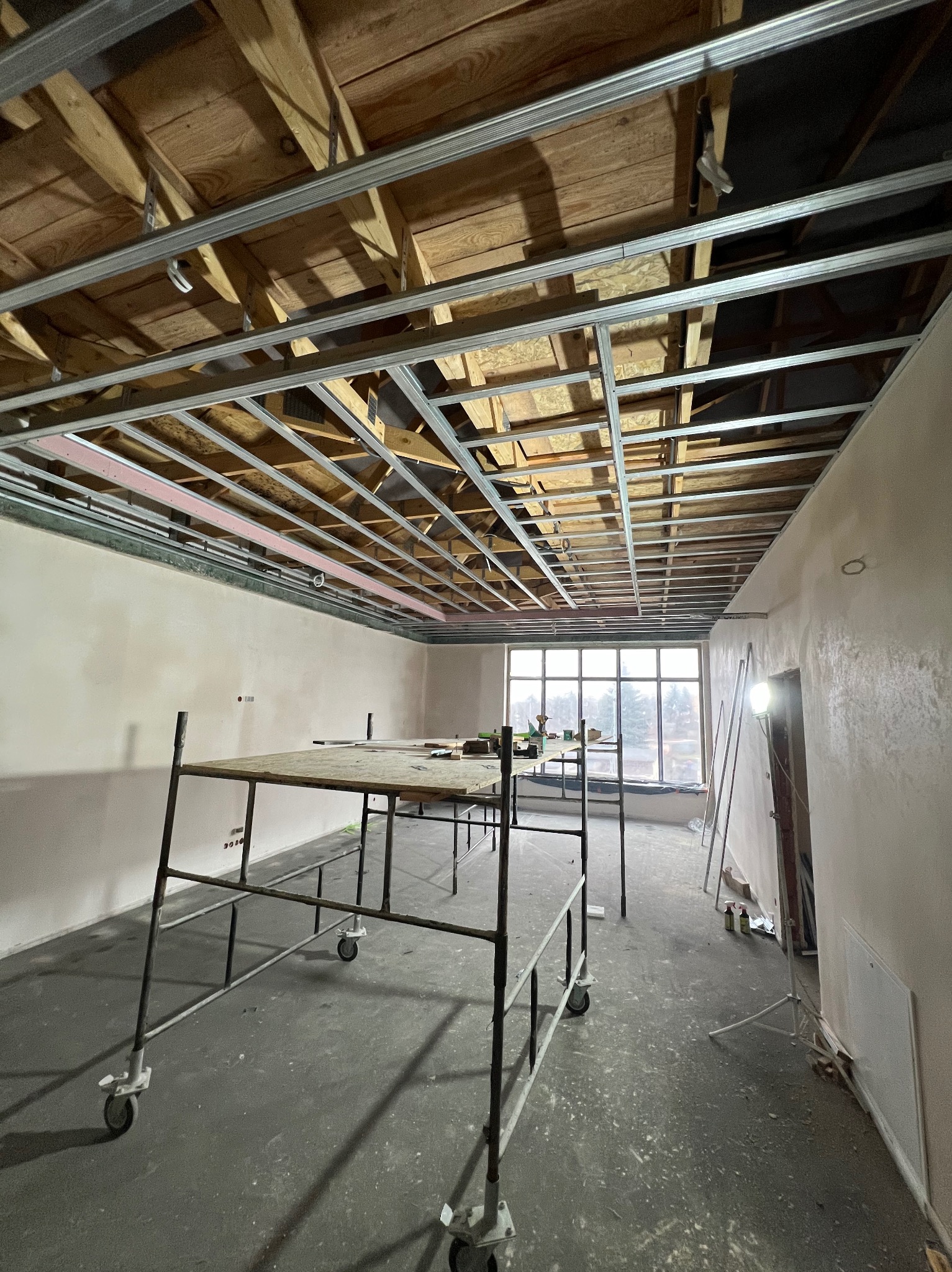 Montaż metalowej konstrukcji sufitu podwieszanego w jasnym pomieszczeniu z oknem. Widoczne rusztowanie i narzędzia. Etap prac wykończeniowych.