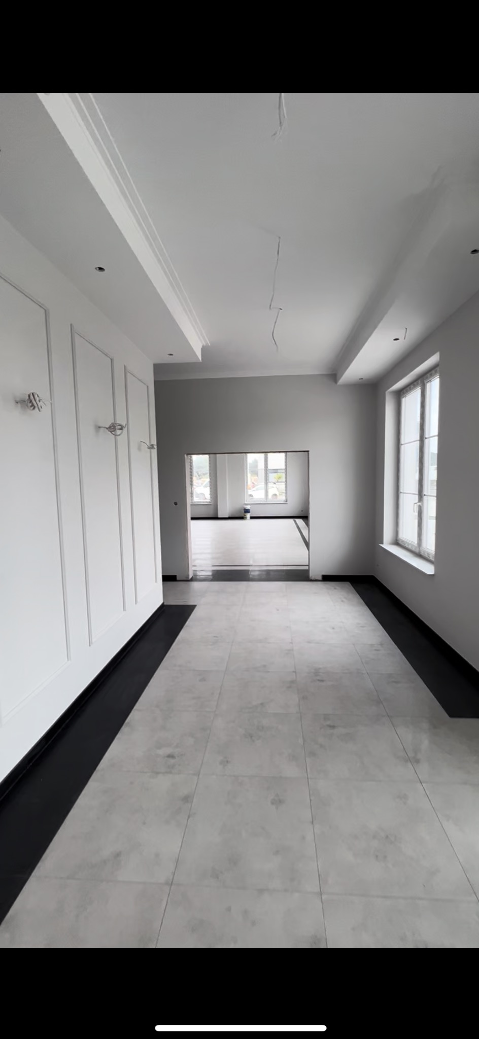 Białe wnętrze z szarą podłogą z płytek, dekoracyjne listwy ścienne i sufitowe, okno z białymi ramami. Widok na kolejne pomieszczenie.