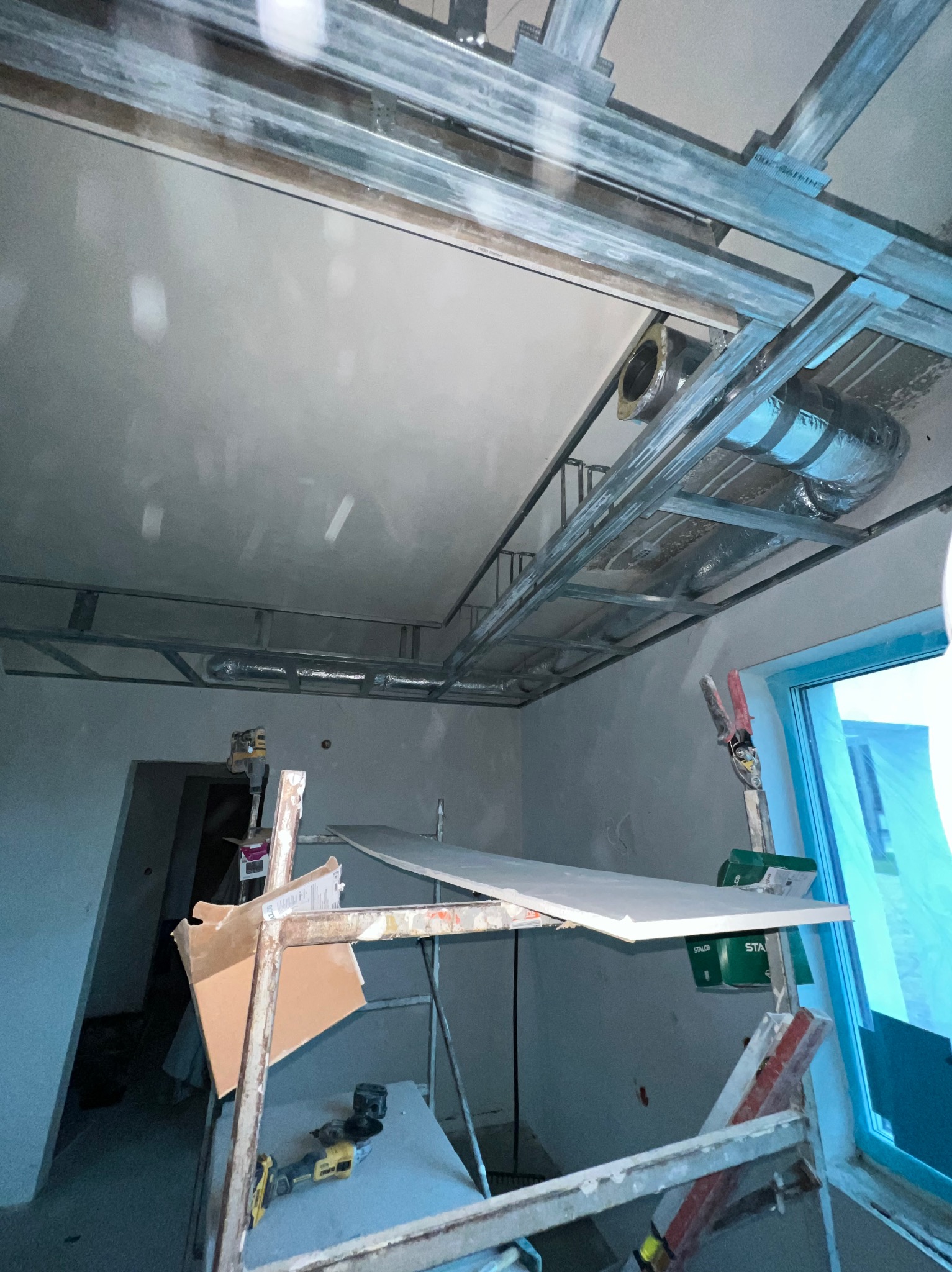 Montaż sufitu podwieszanego z płyt karton-gips na metalowej konstrukcji. Widoczne elementy wentylacji i rusztowanie z narzędziami. Ujęcie z dołu, w trakcie prac.