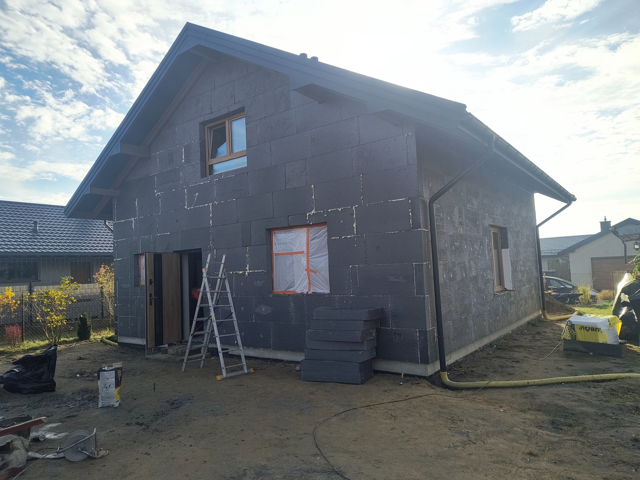 Dom w trakcie ocieplania, ściany pokryte grafitowym styropianem. Widoczne okna, drabina oparta o ścianę, materiały budowlane. Słoneczny dzień, błękitne niebo.