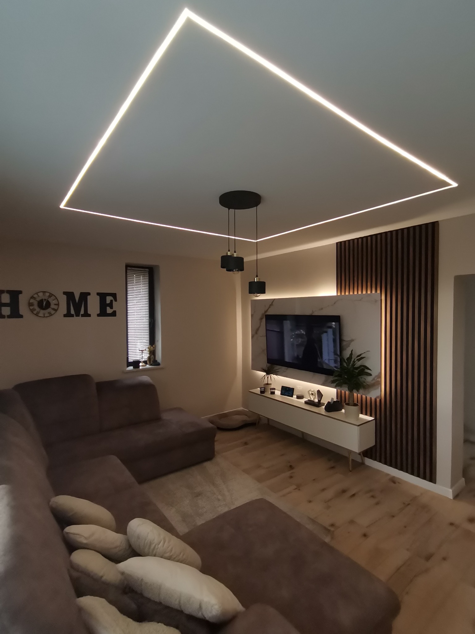 Nowoczesny salon z narożnikiem, drewnianą ścianką TV i podświetleniem LED w suficie. Minimalistyczny design, ciepłe oświetlenie, parkiet na podłodze. Dekoracja HOME na ścianie.