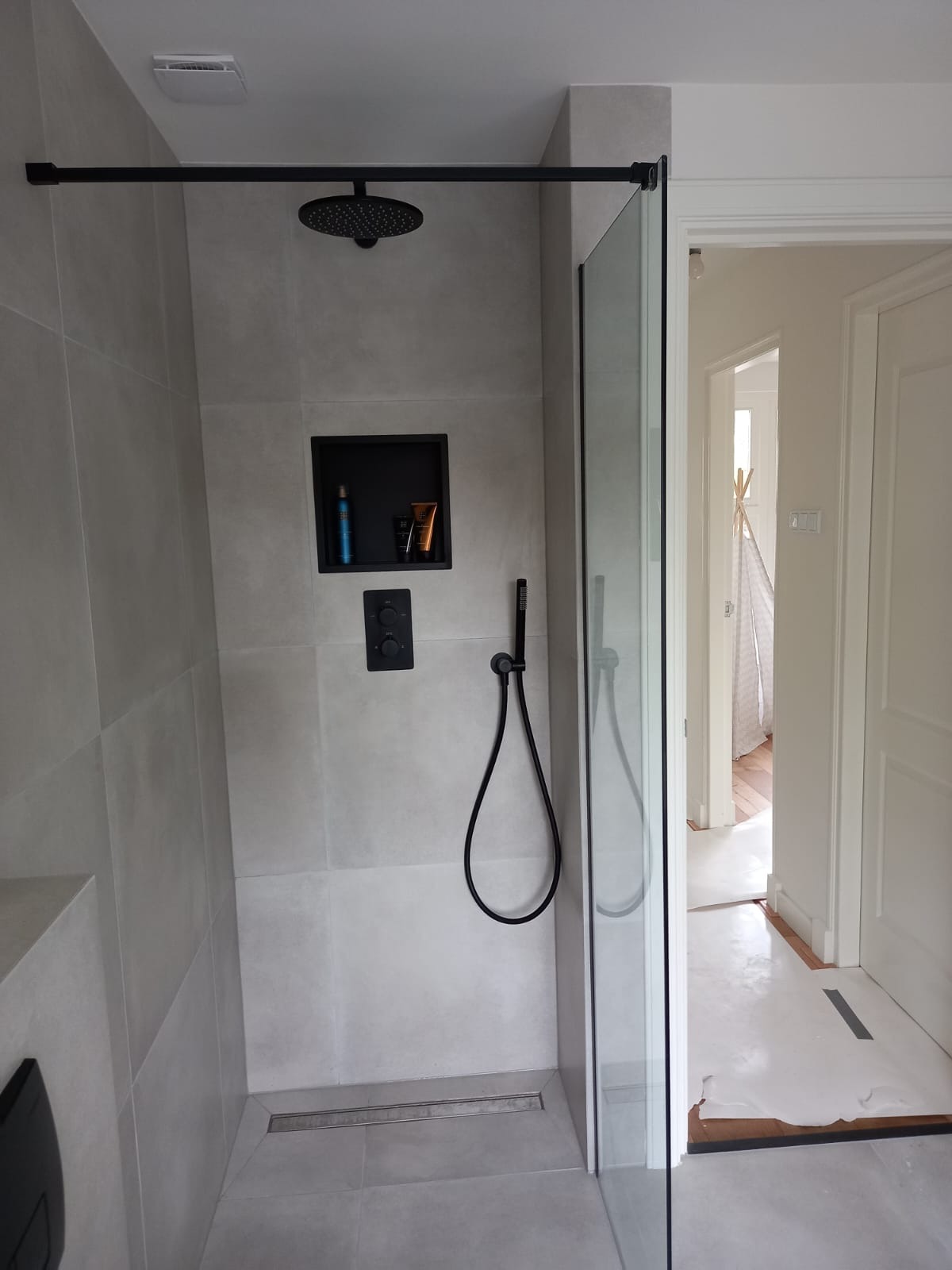 Nowoczesna kabina prysznicowa z szarymi płytkami, czarną armaturą i wnęką na kosmetyki. Minimalistyczny design, widok na wnętrze łazienki przez szklane drzwi.