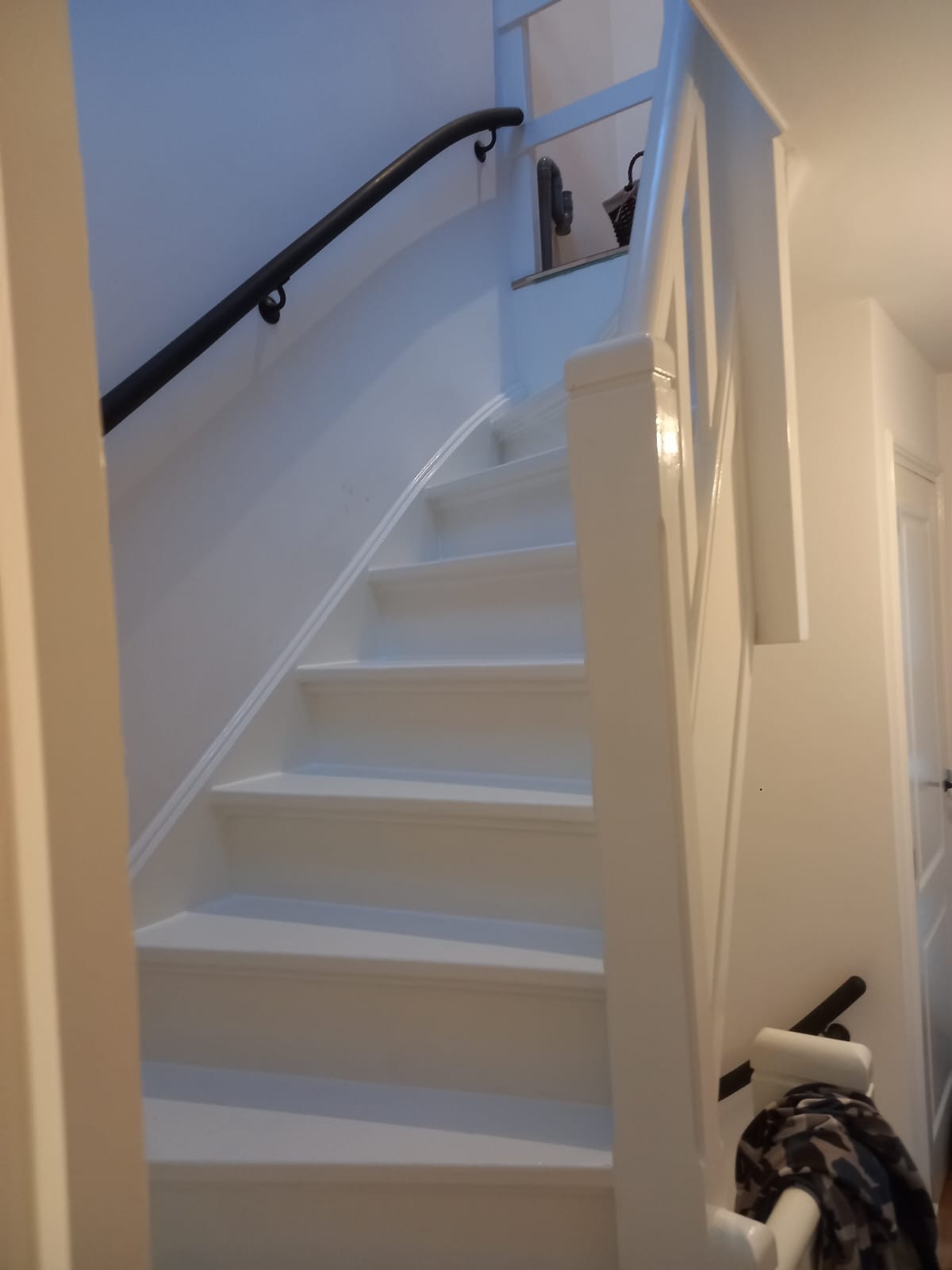 Białe, odnowione schody z czarną poręczą, widok z dołu. Świeżo pomalowane stopnie i barierki, jasne wnętrze. Widoczny fragment drzwi po prawej stronie.