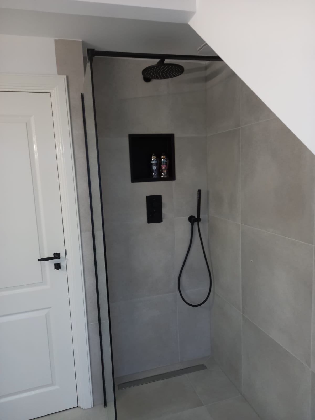 Nowoczesna kabina prysznicowa walk-in z czarnymi elementami: deszczownica, bateria, wnęka na kosmetyki. Szare płytki, białe drzwi. Minimalistyczny design.